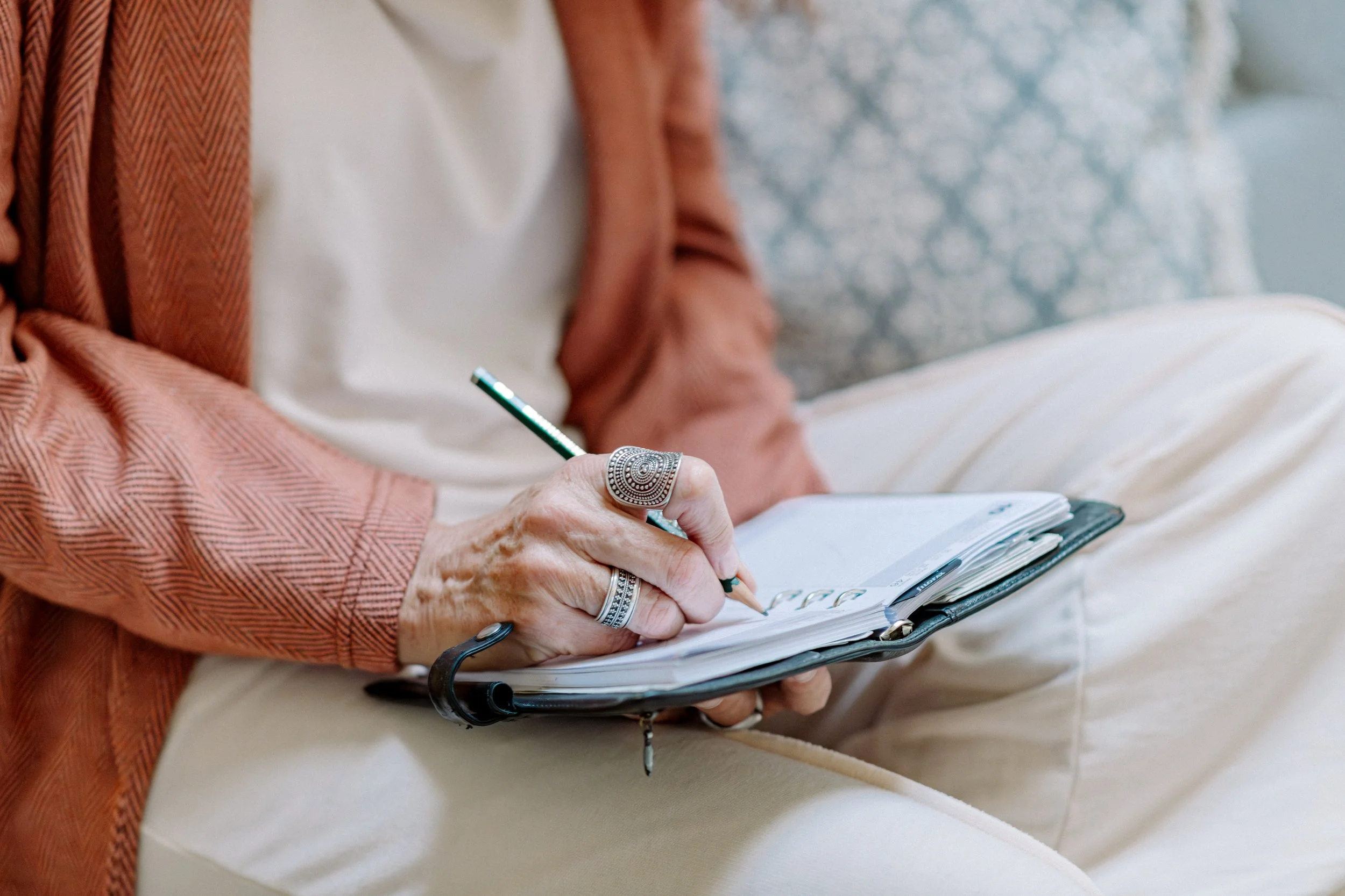 Nahaufnahme einer Person, die in einem Notizbuch schreibt, während sie auf einem Sofa sitzt. Die Person trägt Ringe und hält einen Stift. Der Hintergrund ist unscharf mit einem hellblauen, gemusterten Kissen.