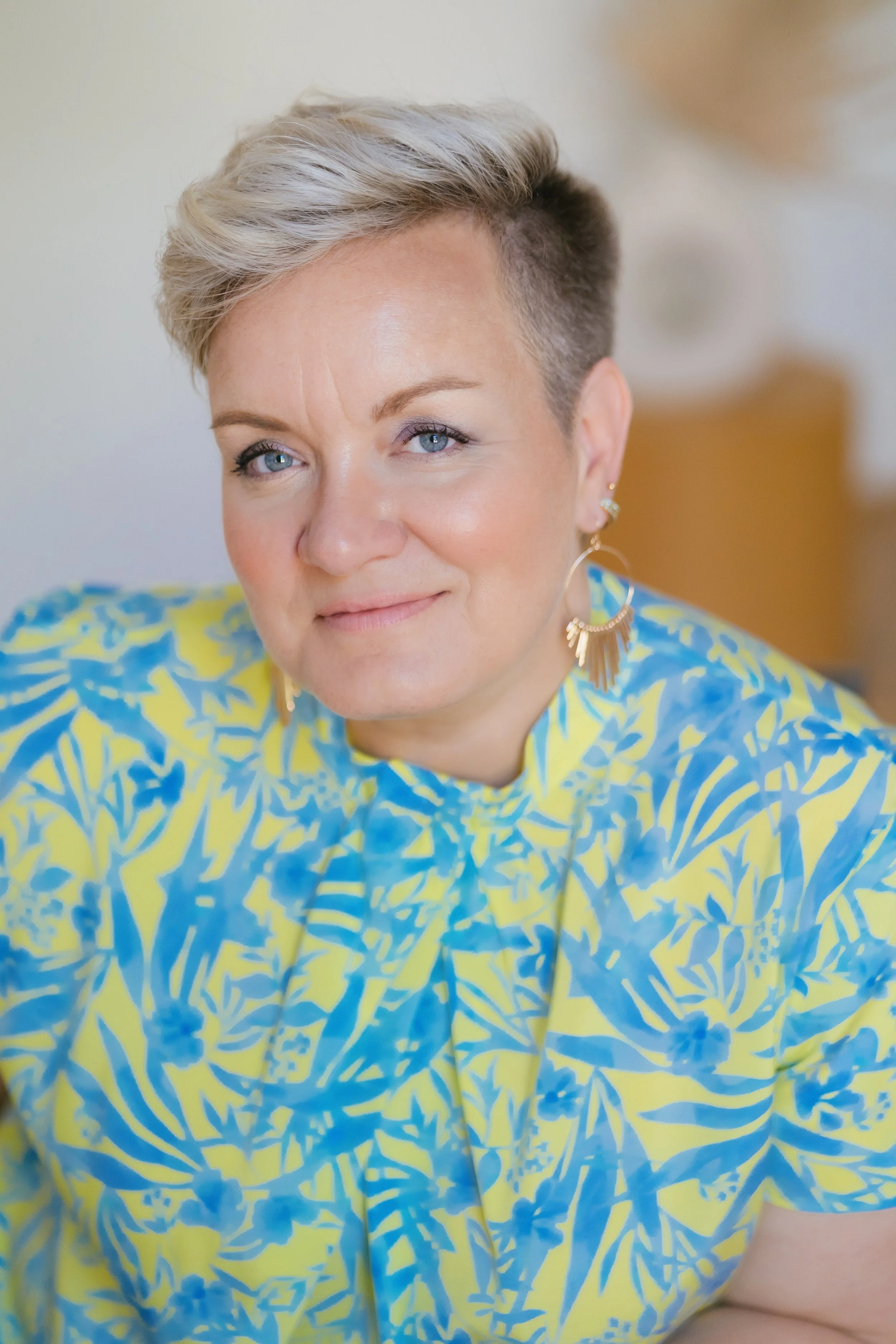 Nahaufnahme eines lachenden Frauengesichts mit kurzen, grauen Haaren, blauen Augen, in einer blumigen, gelb-blauen Bluse, mit großen goldenen Ohrringen.