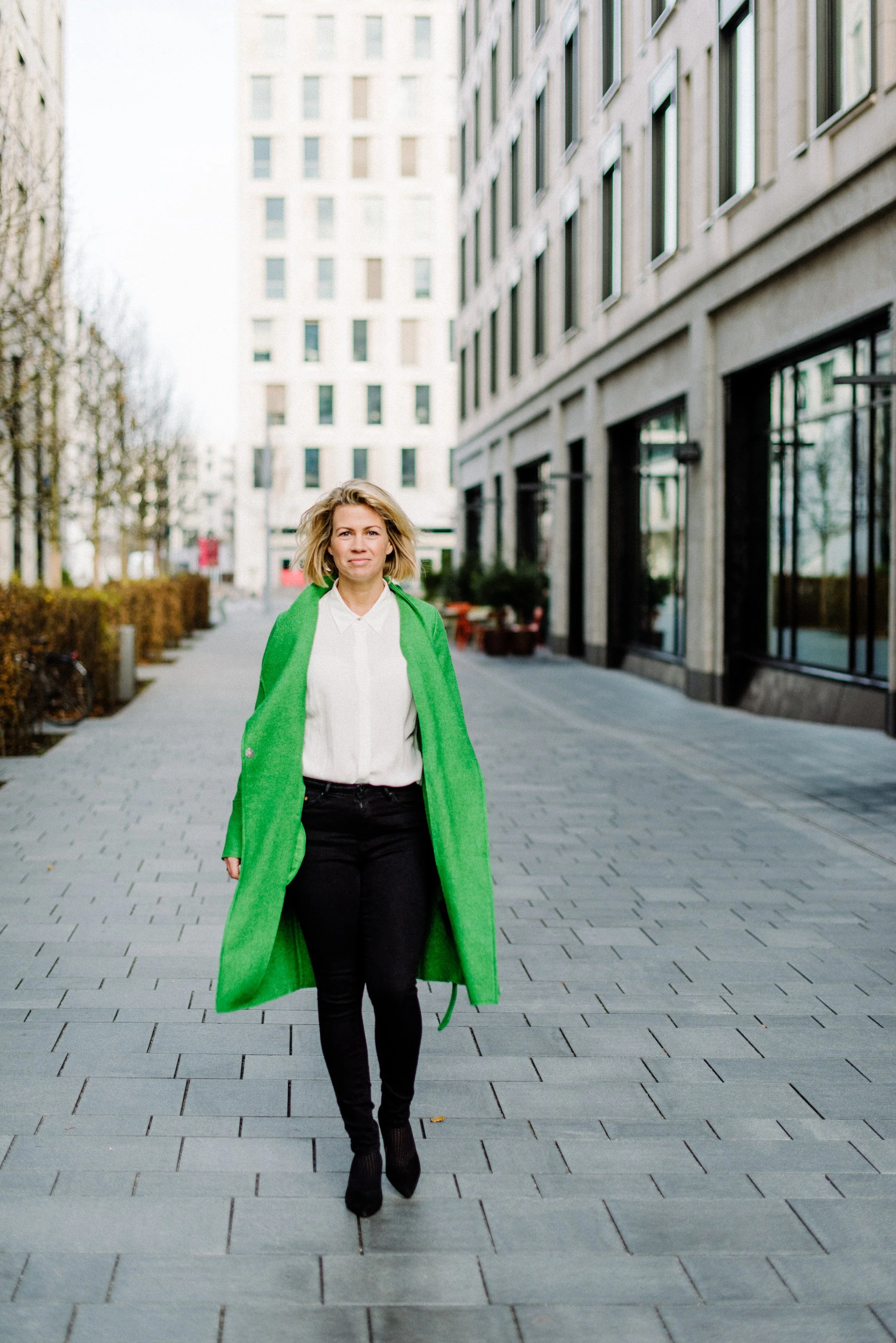 Junge Frau mit blonden Haaren, weißem Hemd und schwarzer Hose, trägt einen grünen Mantel, geht auf einer Straße in einer Stadt, Hintergrund mit modernen Gebäuden.