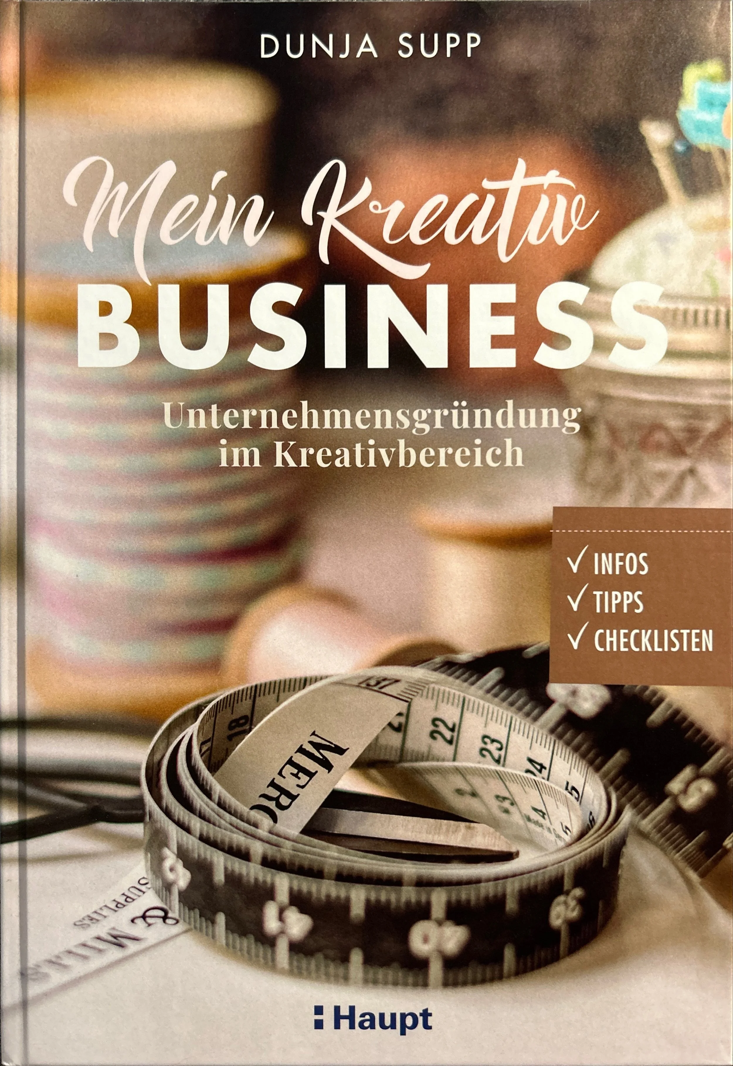 Buchcover mit dem Titel "Mein Kreativ Business" von Dunjá Supp. Das Cover zeigt eine Nahaufnahme eines Maßbands, der zu einer Spirale aufgewickelt ist. Im Hintergrund sind unscharf bunte Cupcakes zu sehen.