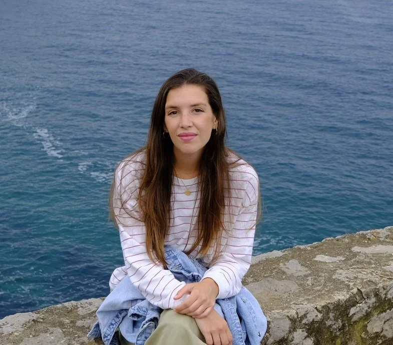Joven mujer sentada en un muro junto al mar, con agua azul de fondo.