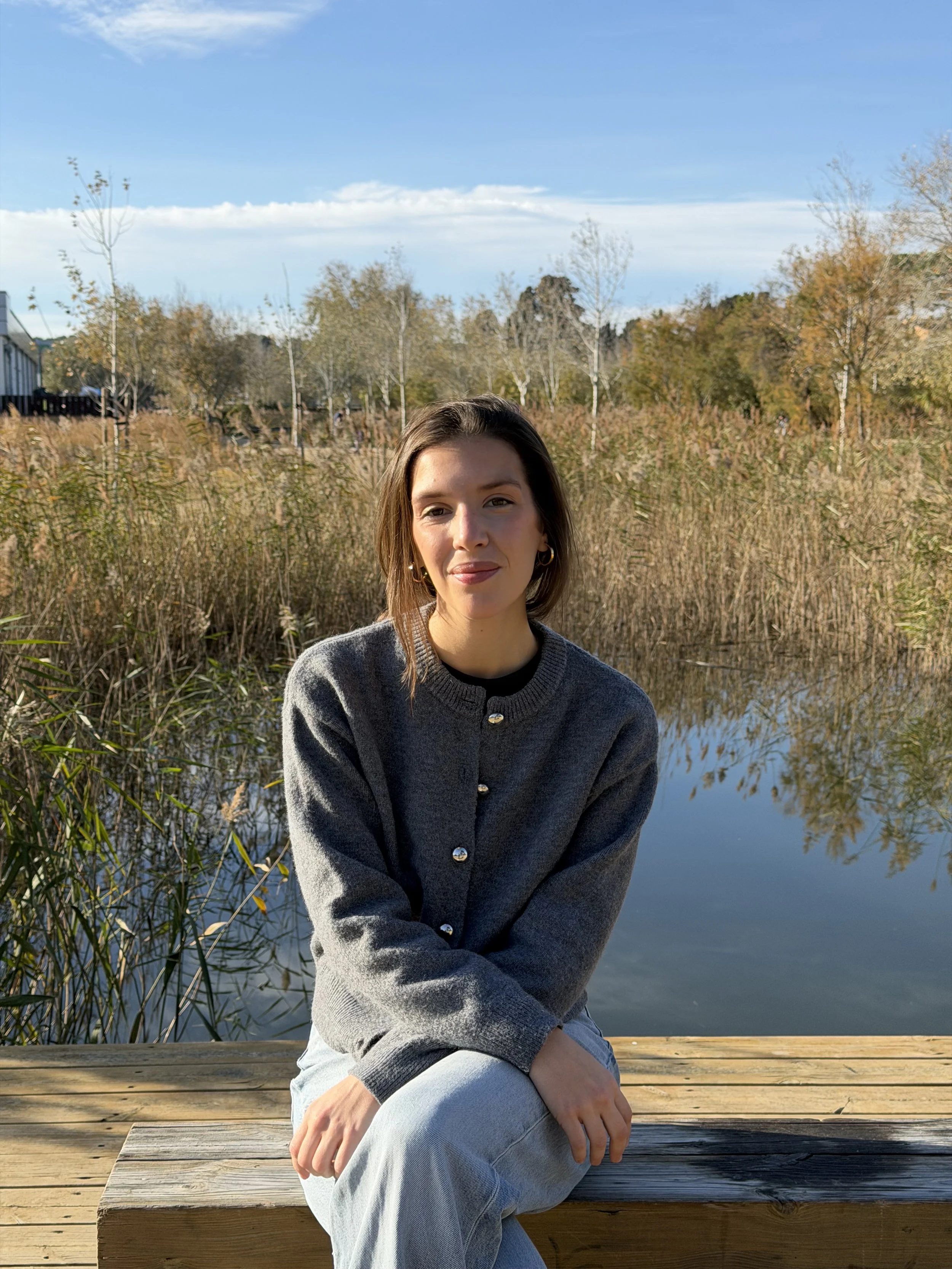 Mujer sentada en un banco de madera junto a un cuerpo de agua, con árboles y cielo despejado de fondo.
