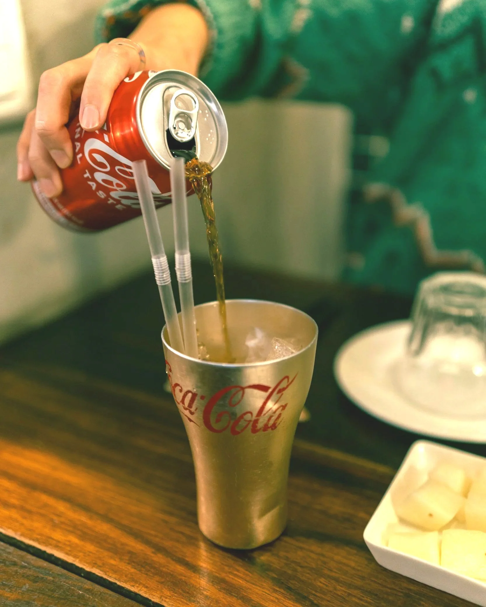 Person pouring Coca-Cola from a soda can into a Coca-Cola branded glass with ice, on a wooden table.