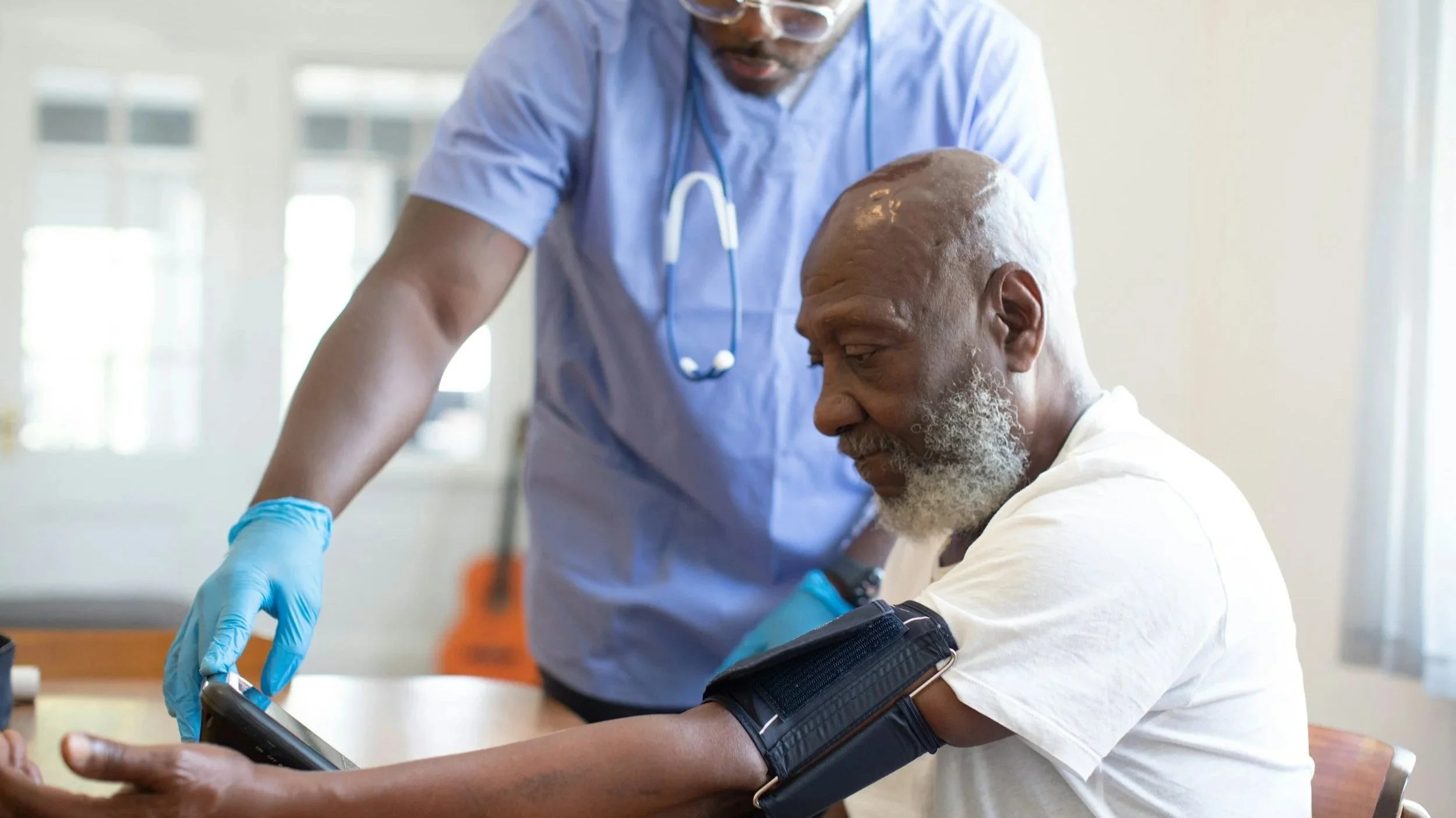 A healthcare professional checks a patient's blood pressure using a digital monitor. The patient is an elderly man with a gray beard, seated at a table, wearing a white t-shirt. The healthcare worker wears blue scrubs, a stethoscope around his neck, and blue gloves.