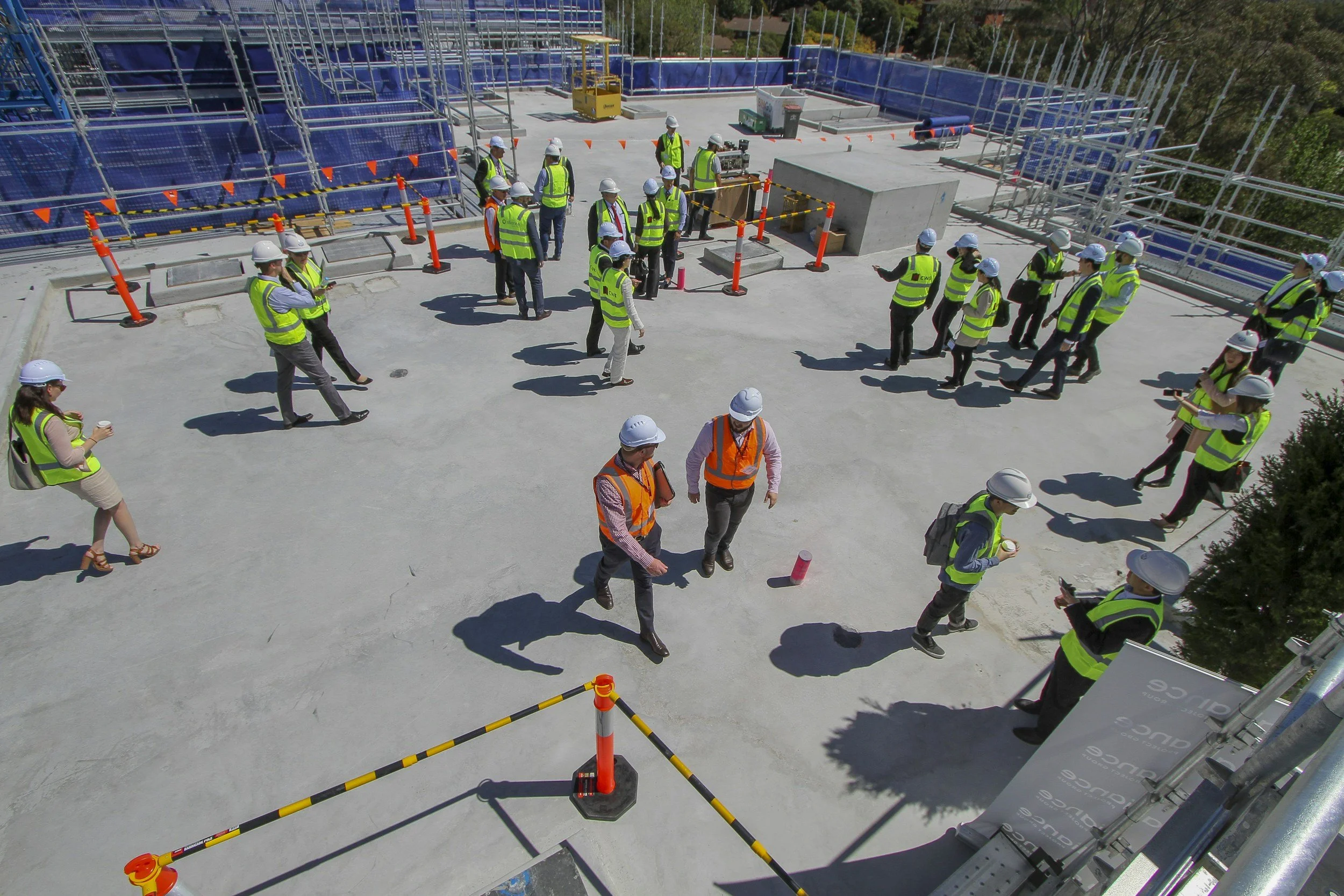 A group of construction workers and visitors, all wearing safety vests and helmets, gathered on a flat concrete rooftop at a construction site, with some scaffolding underneath and trees in the background.