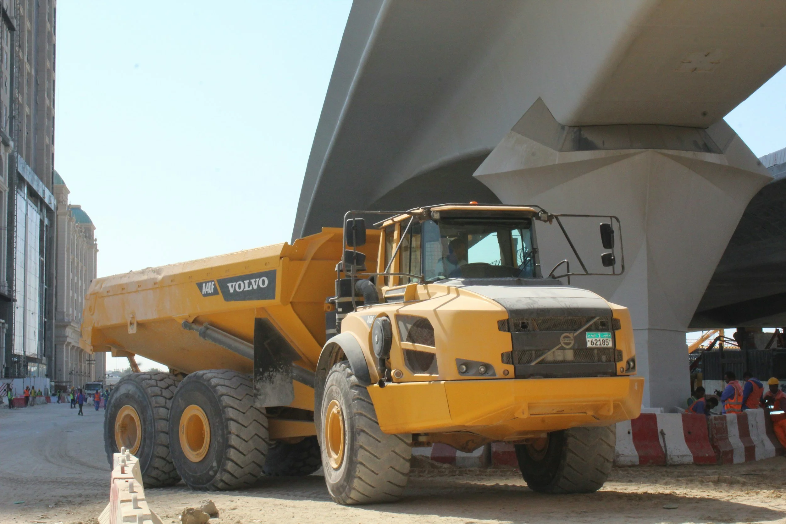 Yellow Volvo construction dump truck parked under a large concrete bridge with construction workers nearby