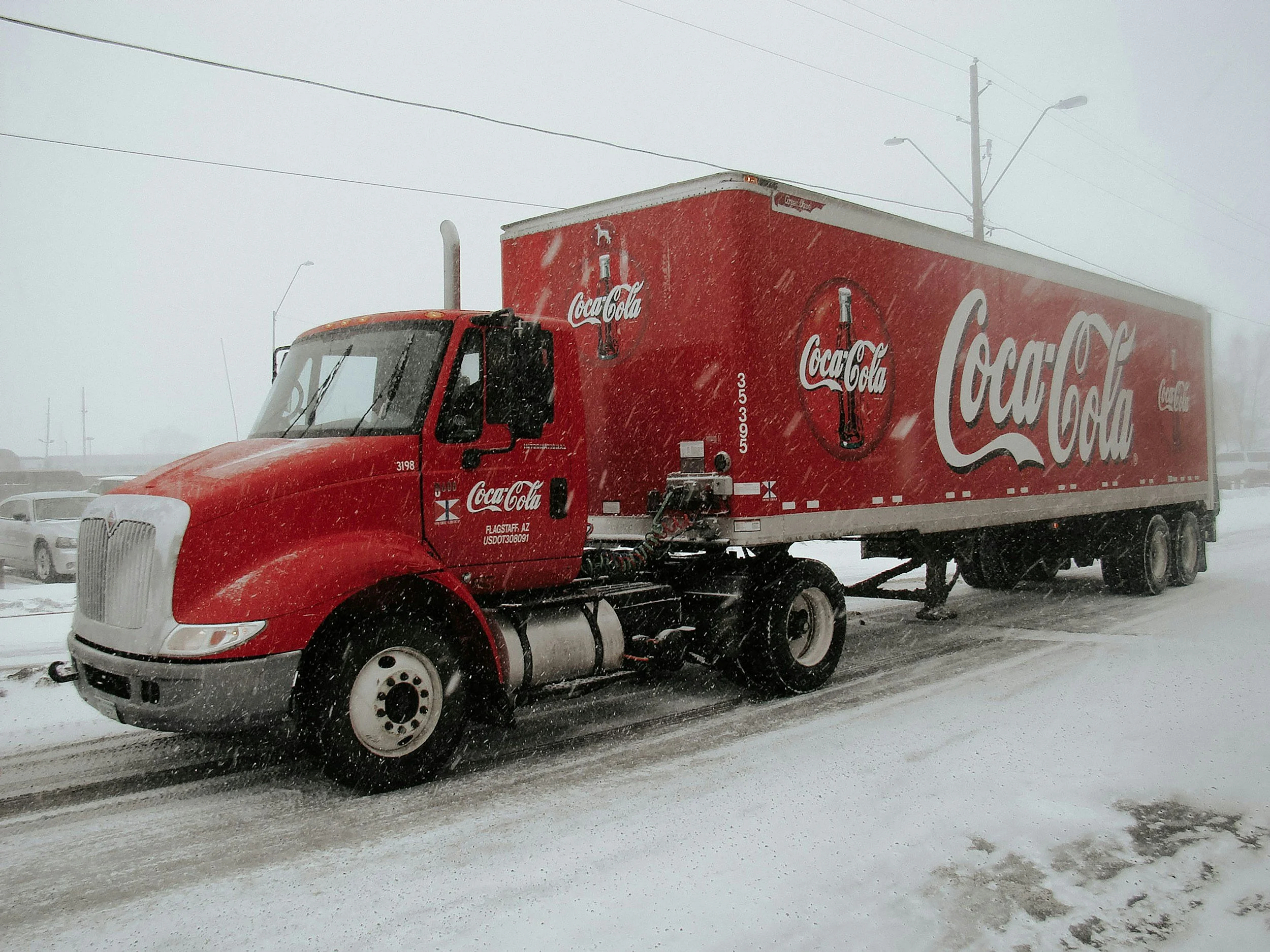 A red Coca-Cola delivery truck parked on a snowy street during a snowstorm, with snow falling heavily and snow-covered ground.
