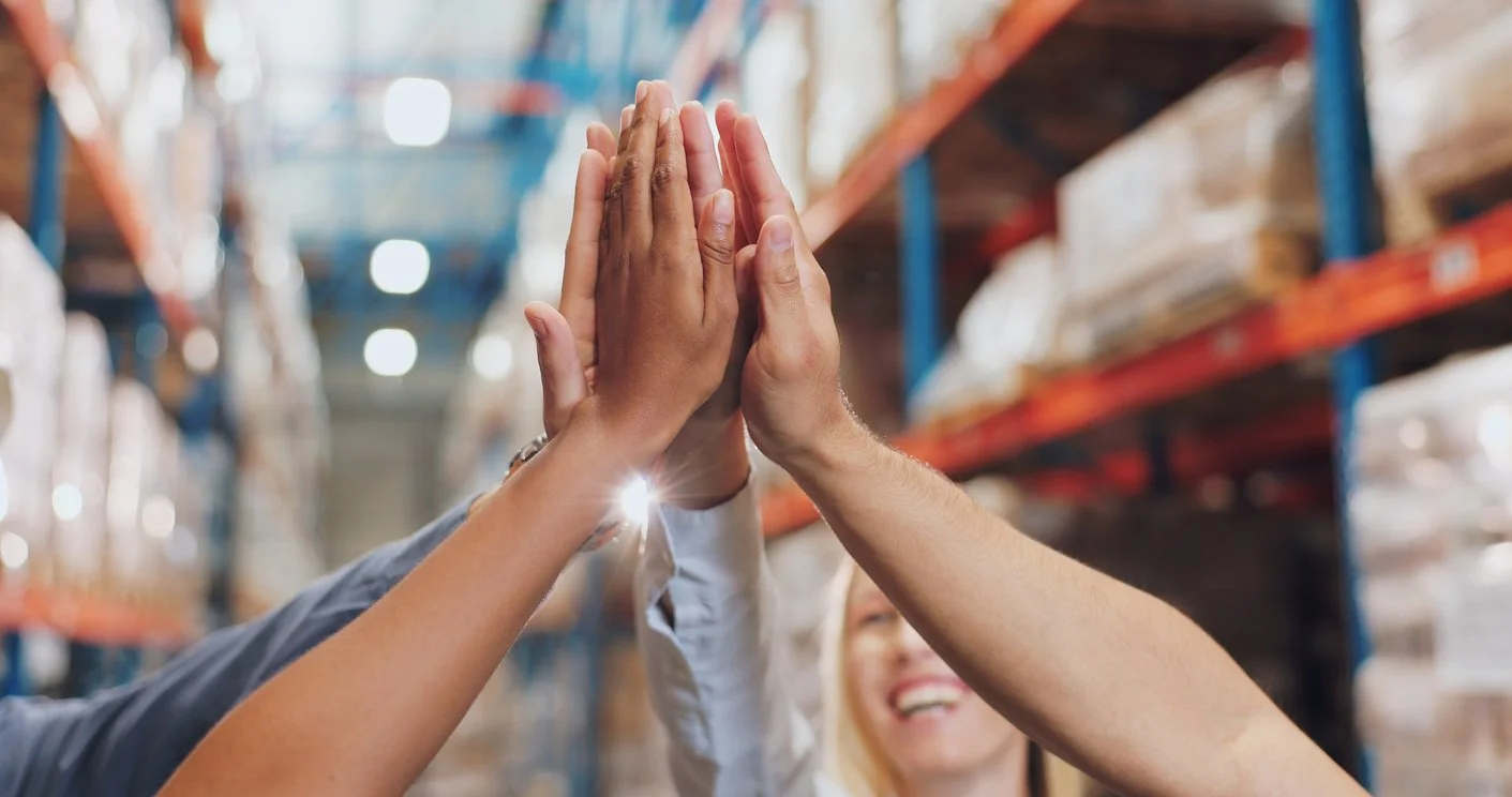 Two pairs of hands high-fiving in a warehouse with shelves of boxes in the background.