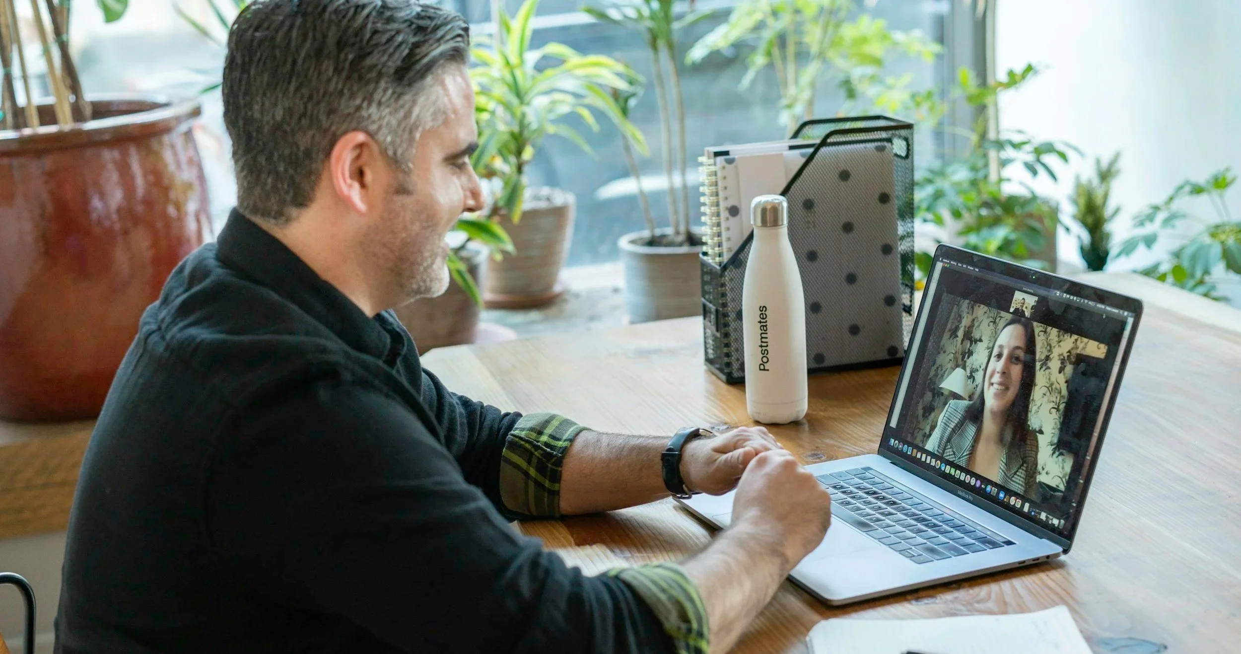A man with gray hair and a dark shirt smiling during a video call with a woman who has dark hair and is wearing a plaid blazer, on a laptop in a bright room with plants and office supplies on the table.