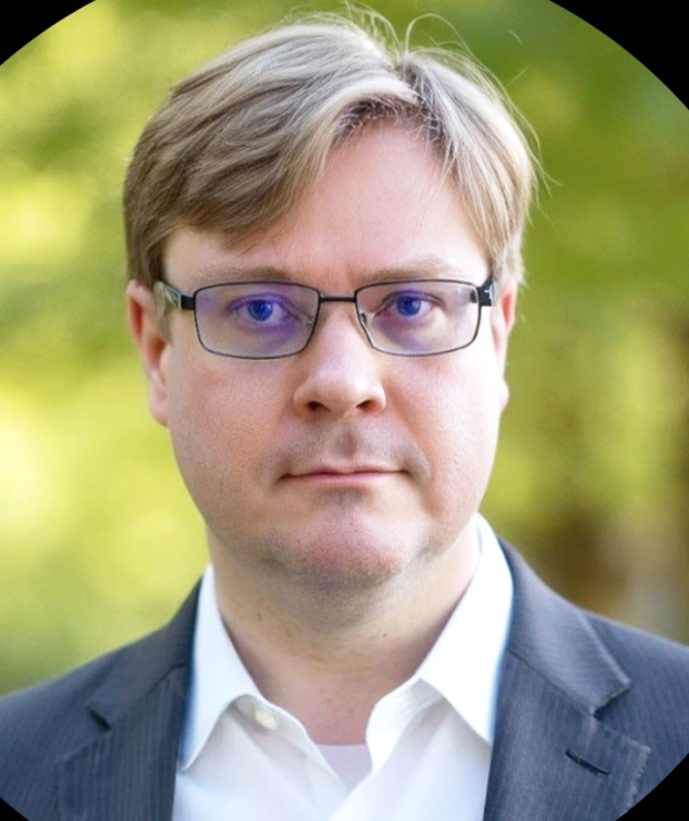 A man wearing glasses and a suit outdoors with green blurred foliage in the background.