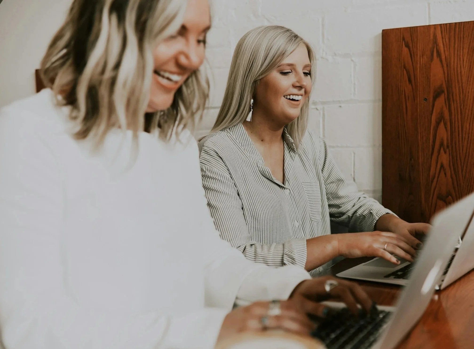 Two women working on laptops, smiling and engaged in a discussion, sitting at a table with a white brick wall background.