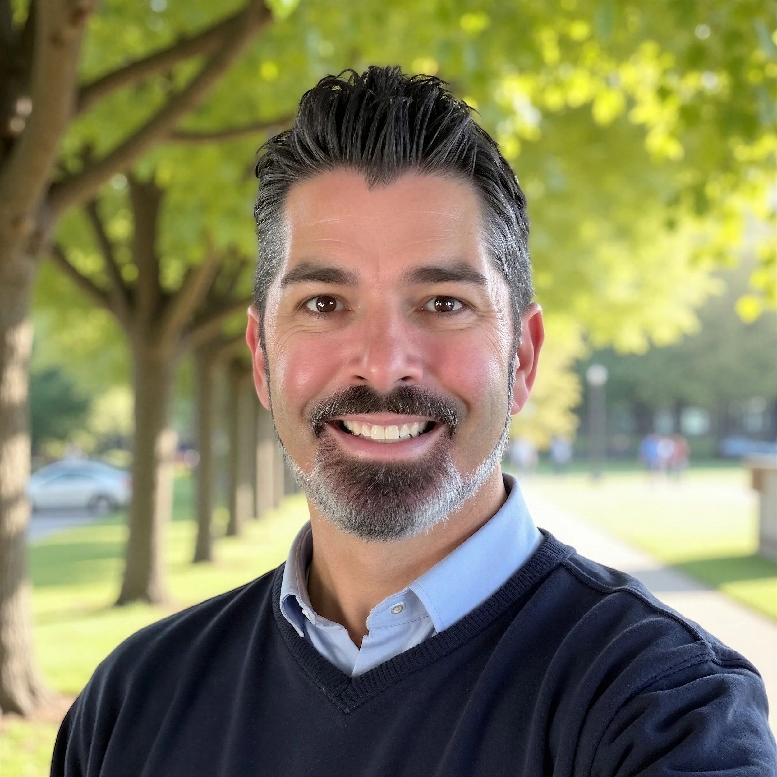 A smiling man with dark hair, a beard, and a mustache, wearing a light blue collared shirt and a dark sweater, standing outdoors in a park with trees and a sidewalk.