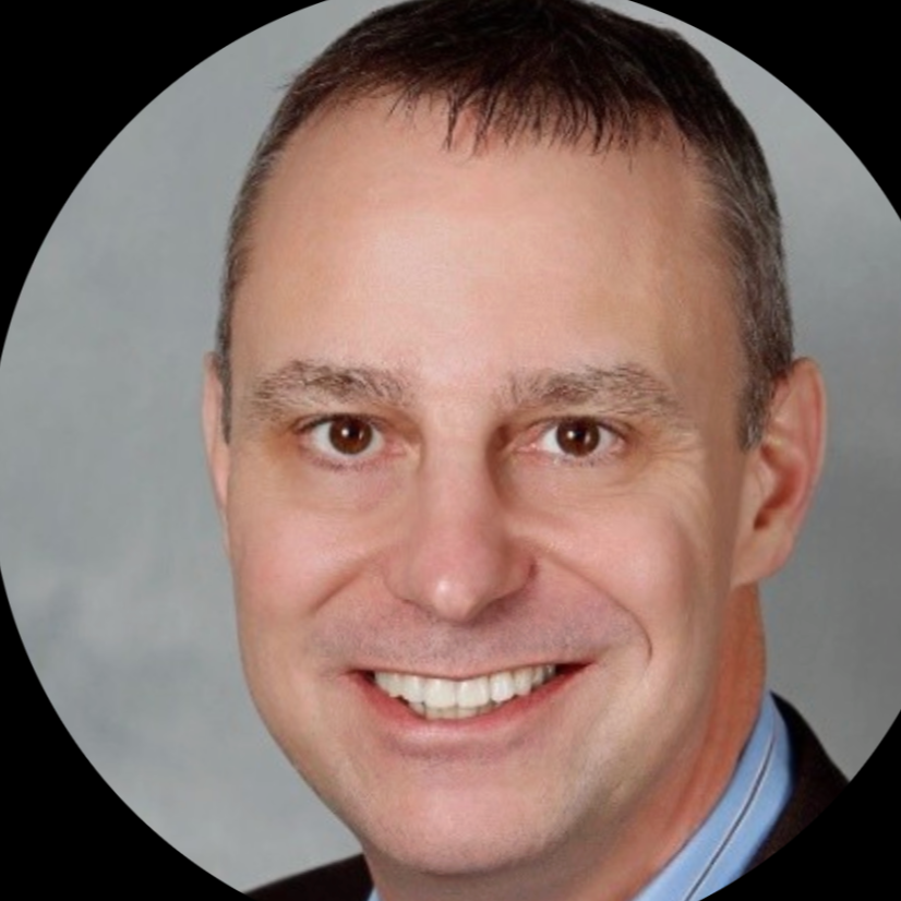 Close-up portrait of a smiling man with short dark hair, light skin, and wearing a suit and tie, against a gray background.