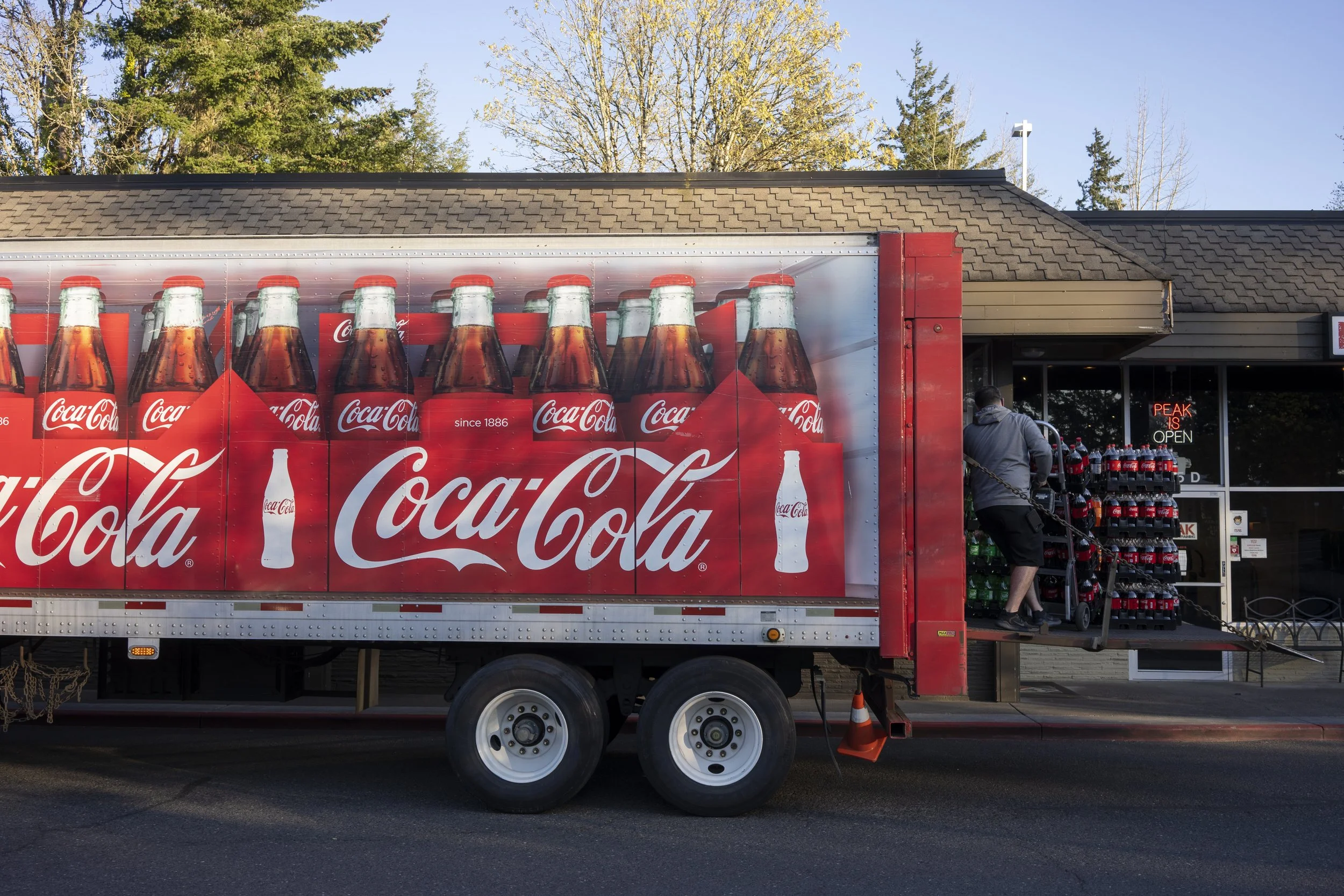 A delivery truck featuring Coca-Cola branding is parked outside a store, with a man unloading soda bottles from the truck and stacking them near the entrance, which has a neon sign that reads "Peak is Open."