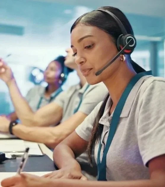 Close-up of a woman wearing a headset with microphone, working at a call center, with two other women in background also wearing headsets and uniforms.