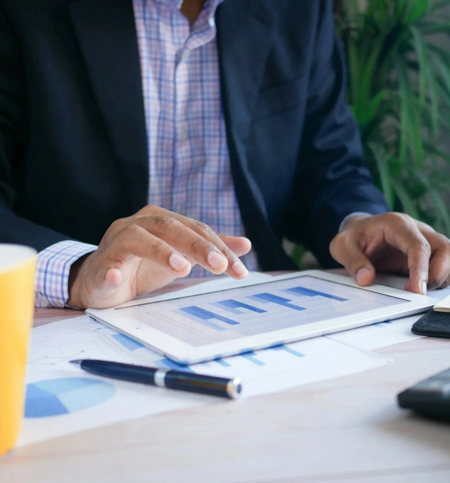 A person in a business suit sitting at a desk using a tablet with graphs and charts, with papers, pen, and other office supplies around.