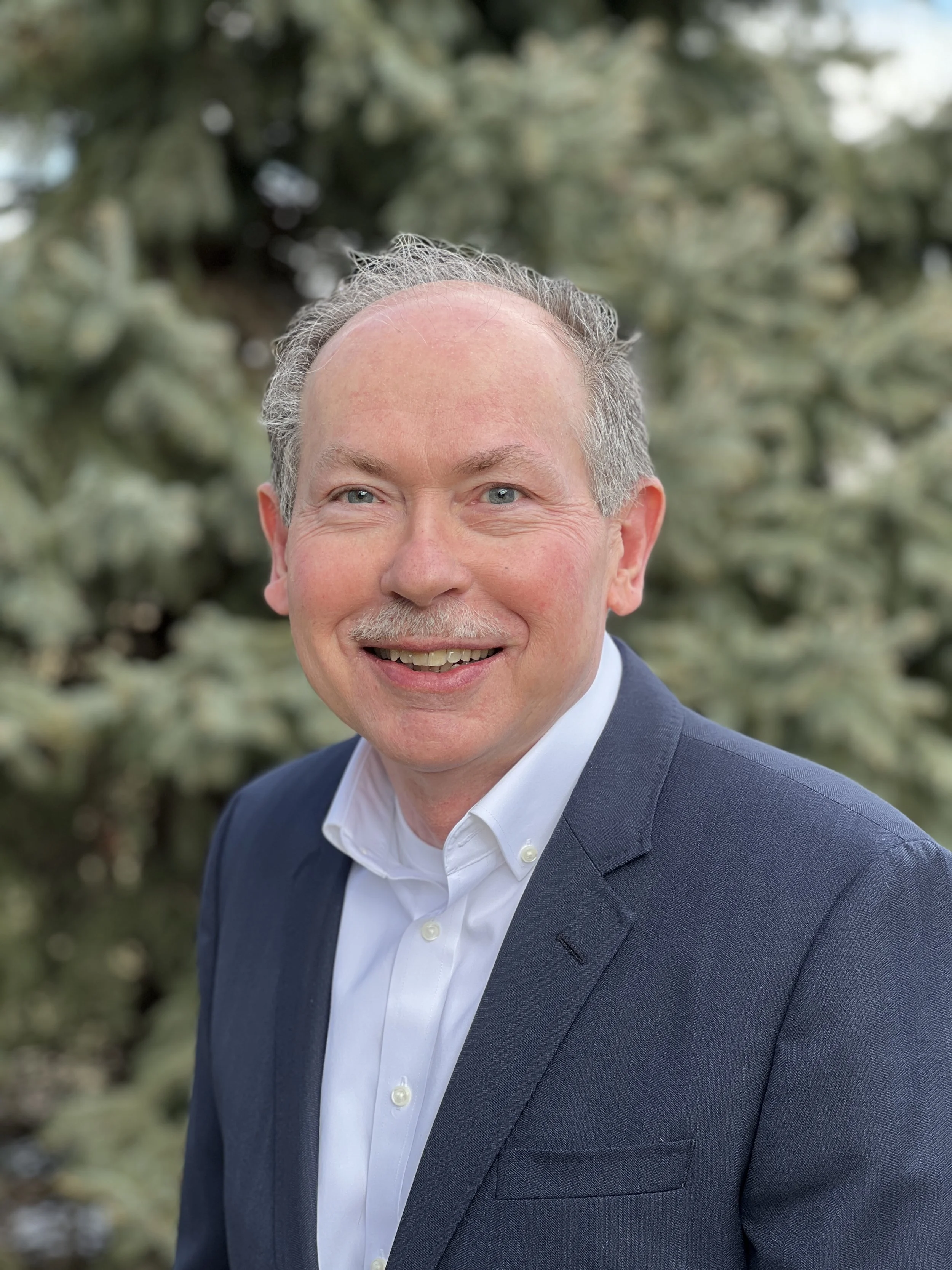 A smiling middle-aged man with blue eyes, gray hair, and a mustache wearing a white dress shirt and dark suit jacket outdoors.