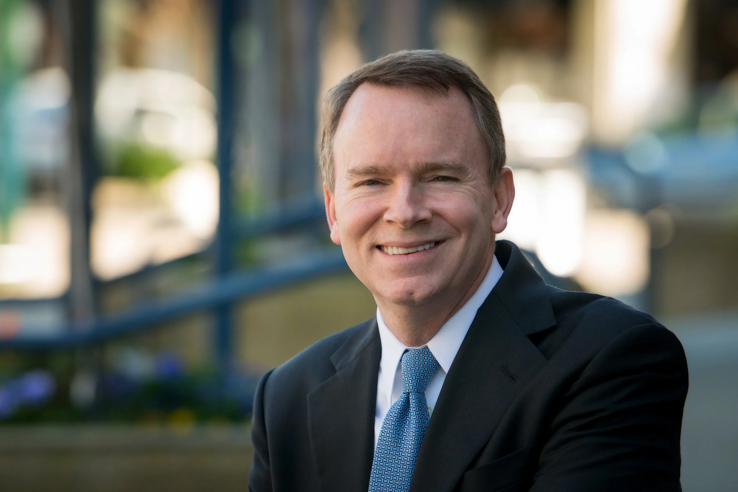 A man in a dark suit and blue tie smiling outdoors with blurred background.