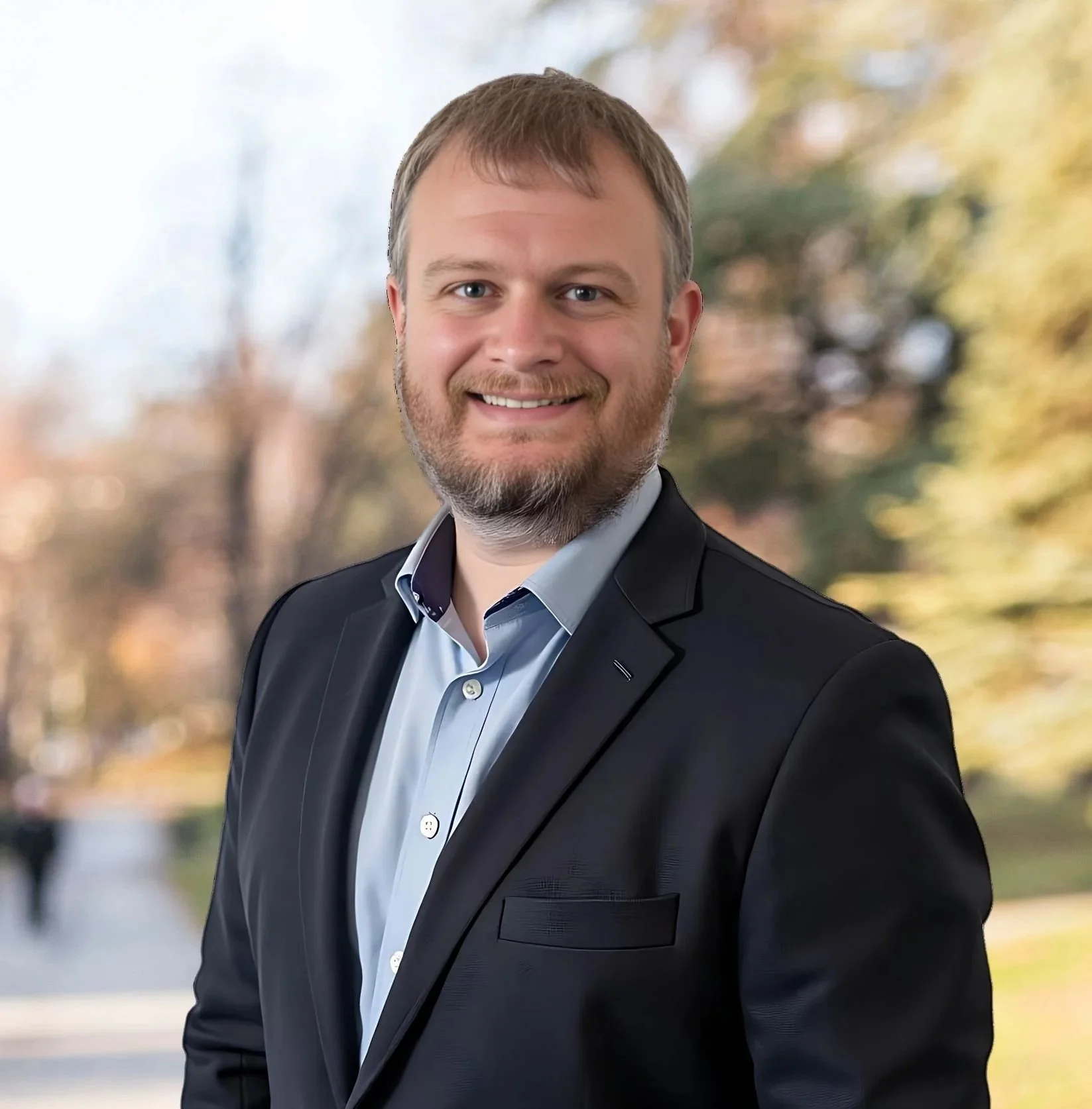 A man in a black suit jacket and light blue shirt smiling outdoors with autumn trees in the background.