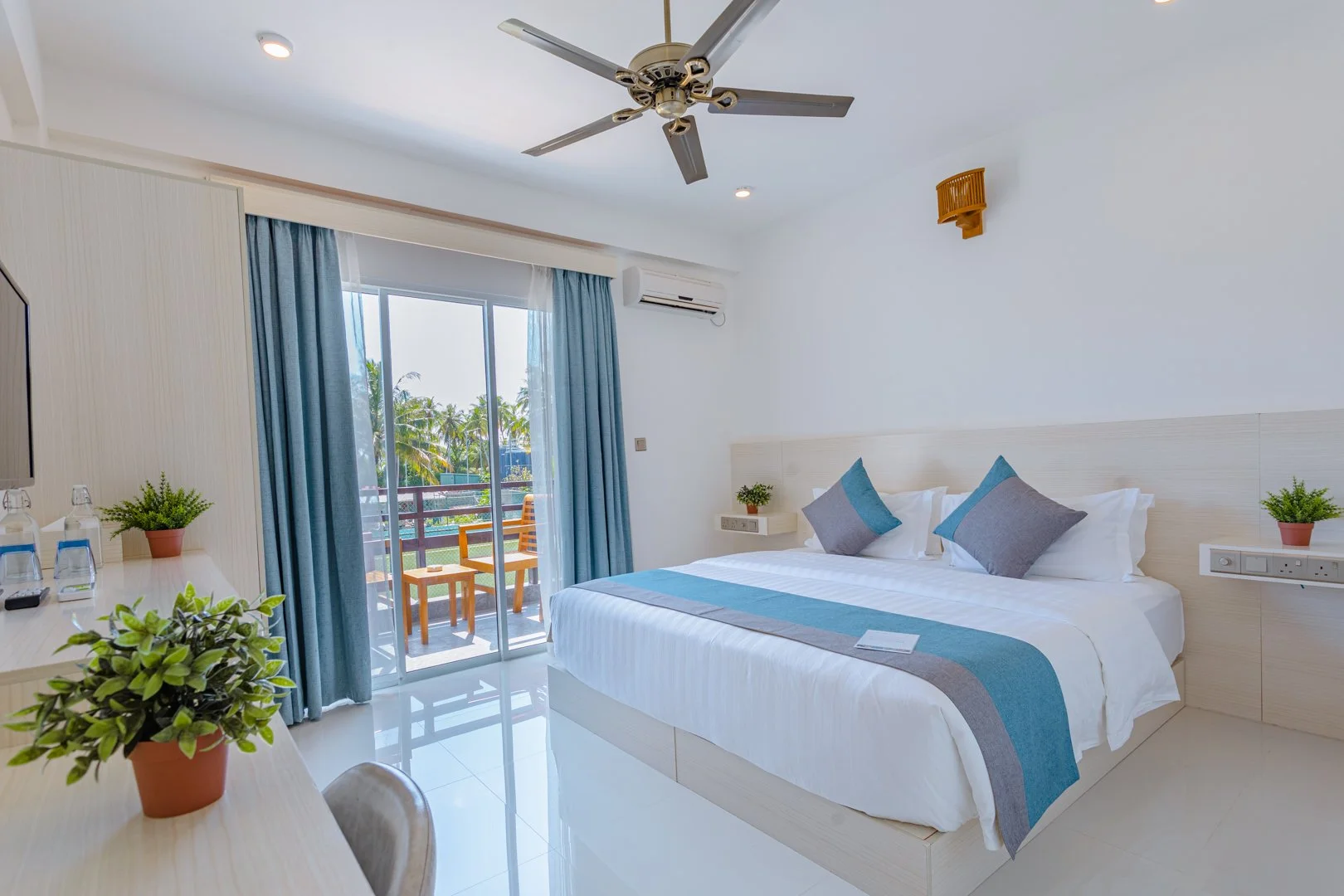 Bright hotel room with a large bed, blue and gray accent pillows, and a sliding glass door opening to a balcony with outdoor seating and tropical trees in the background.