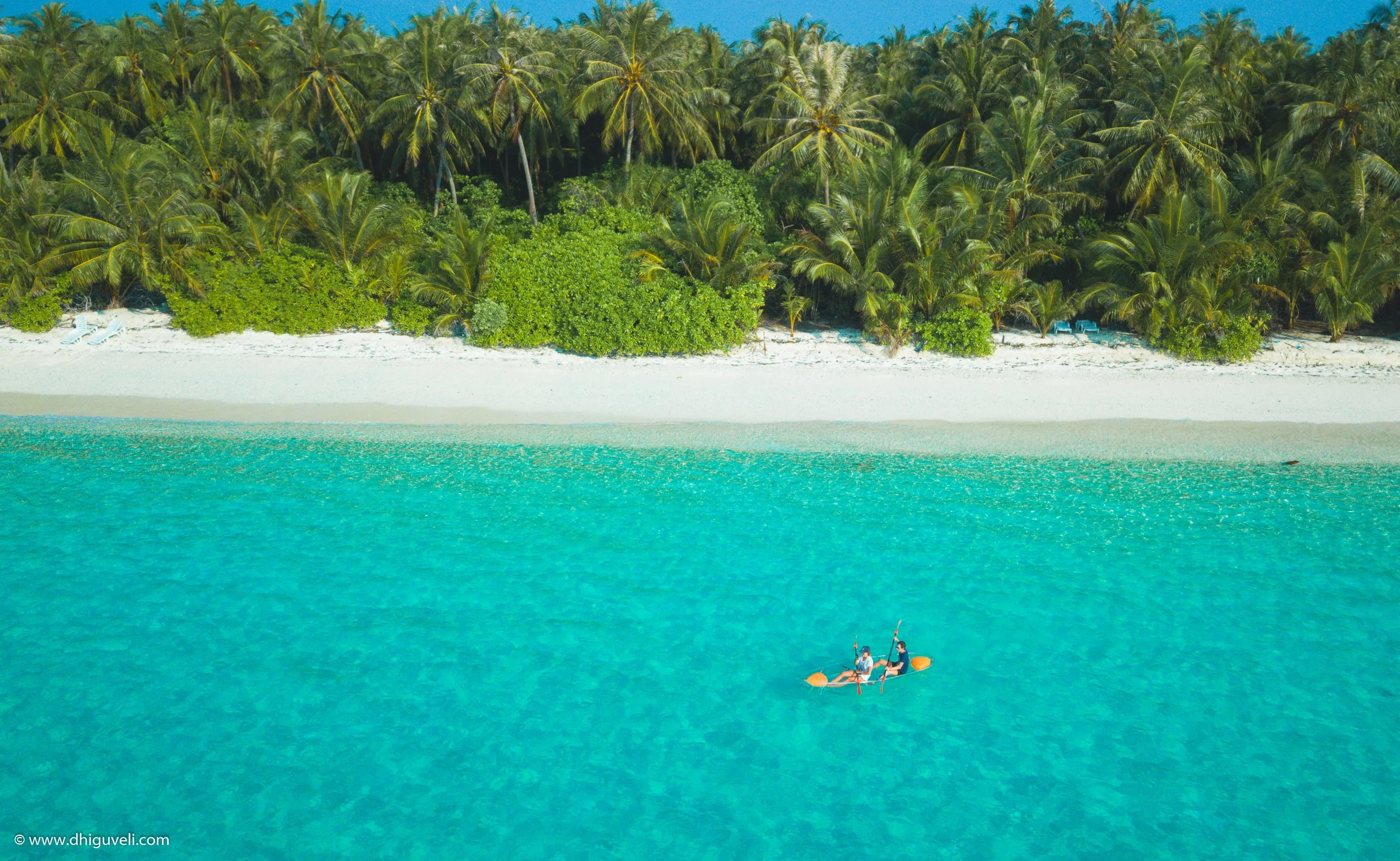 Two people kayaking on clear turquoise water near a white sandy beach and a lush green tropical forest with palm trees.