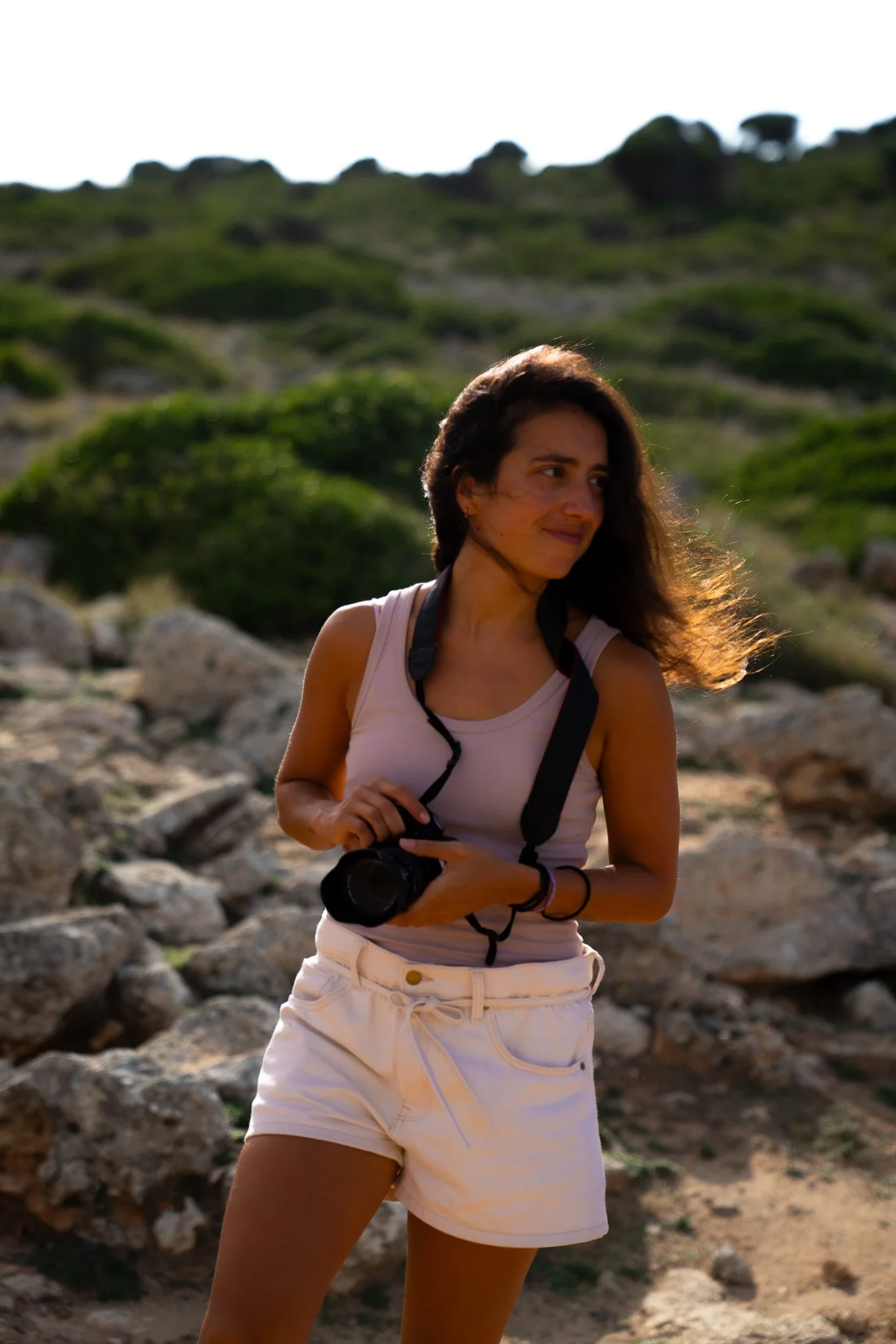 A woman holding a camera outdoors amidst rocky terrain and greenery, with wind-blown hair and sunlight illuminating her face.