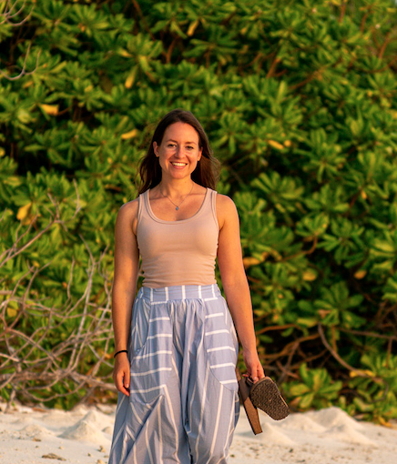 Woman standing outdoors on beach with green foliage in background, smiling and holding a straw hat.