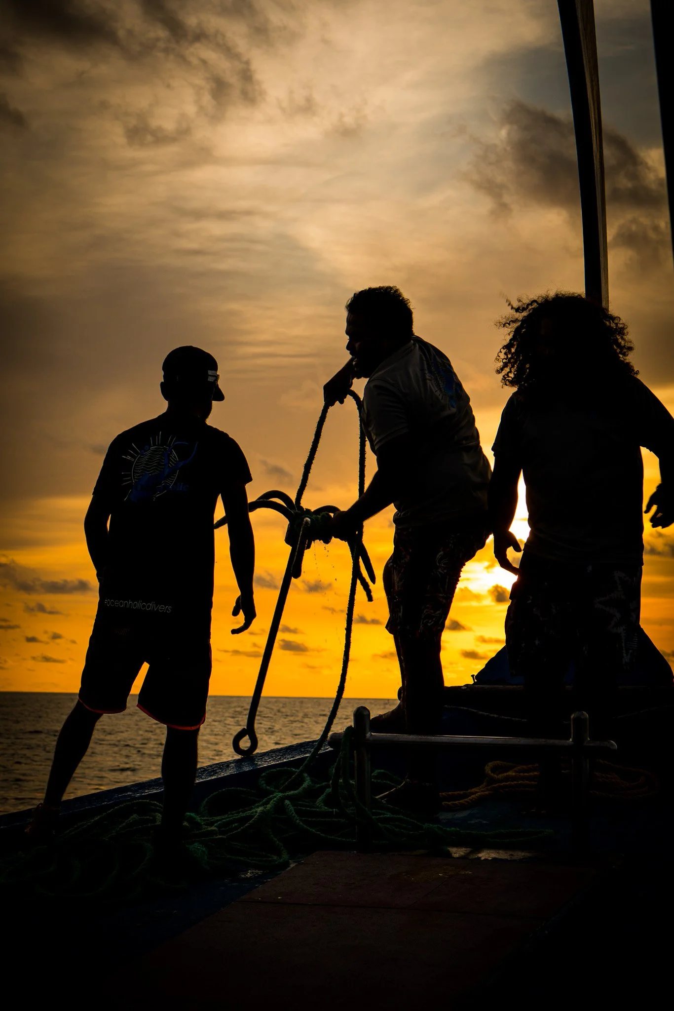 Silhouettes of three people on a boat at sunset, with water and cloudy sky in the background, engaging in a task involving ropes.