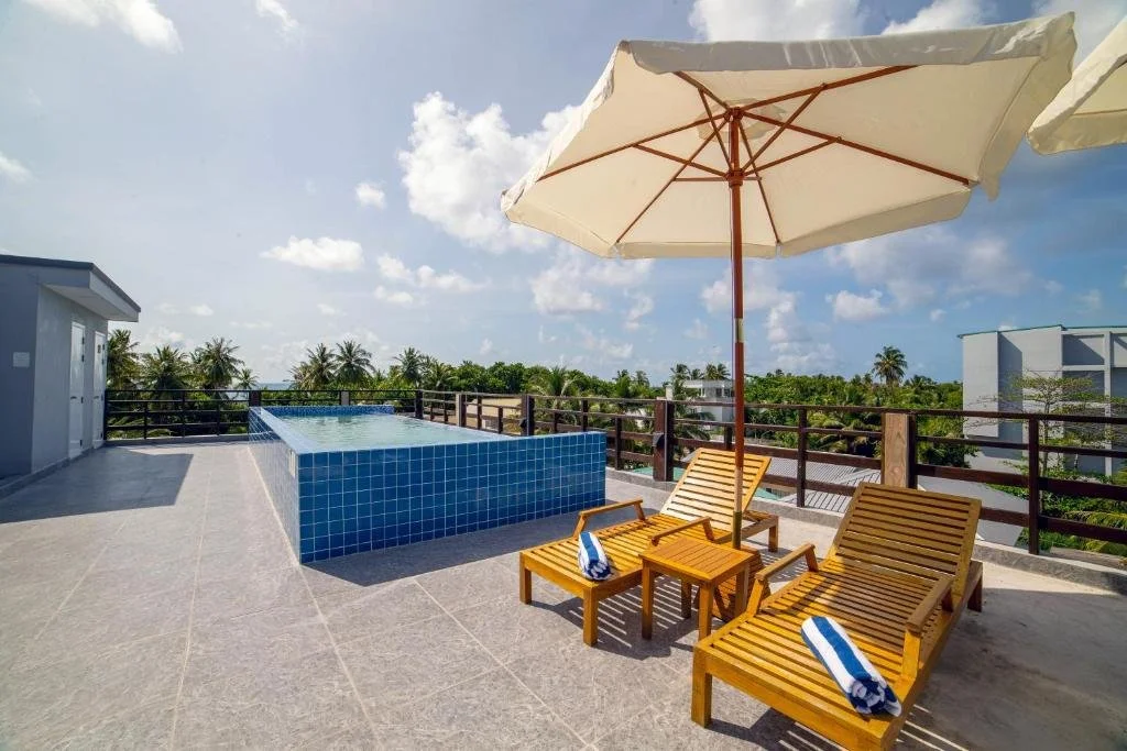 Rooftop pool area with two wooden lounge chairs, an umbrella, and a small side table, overlooking a tropical landscape.