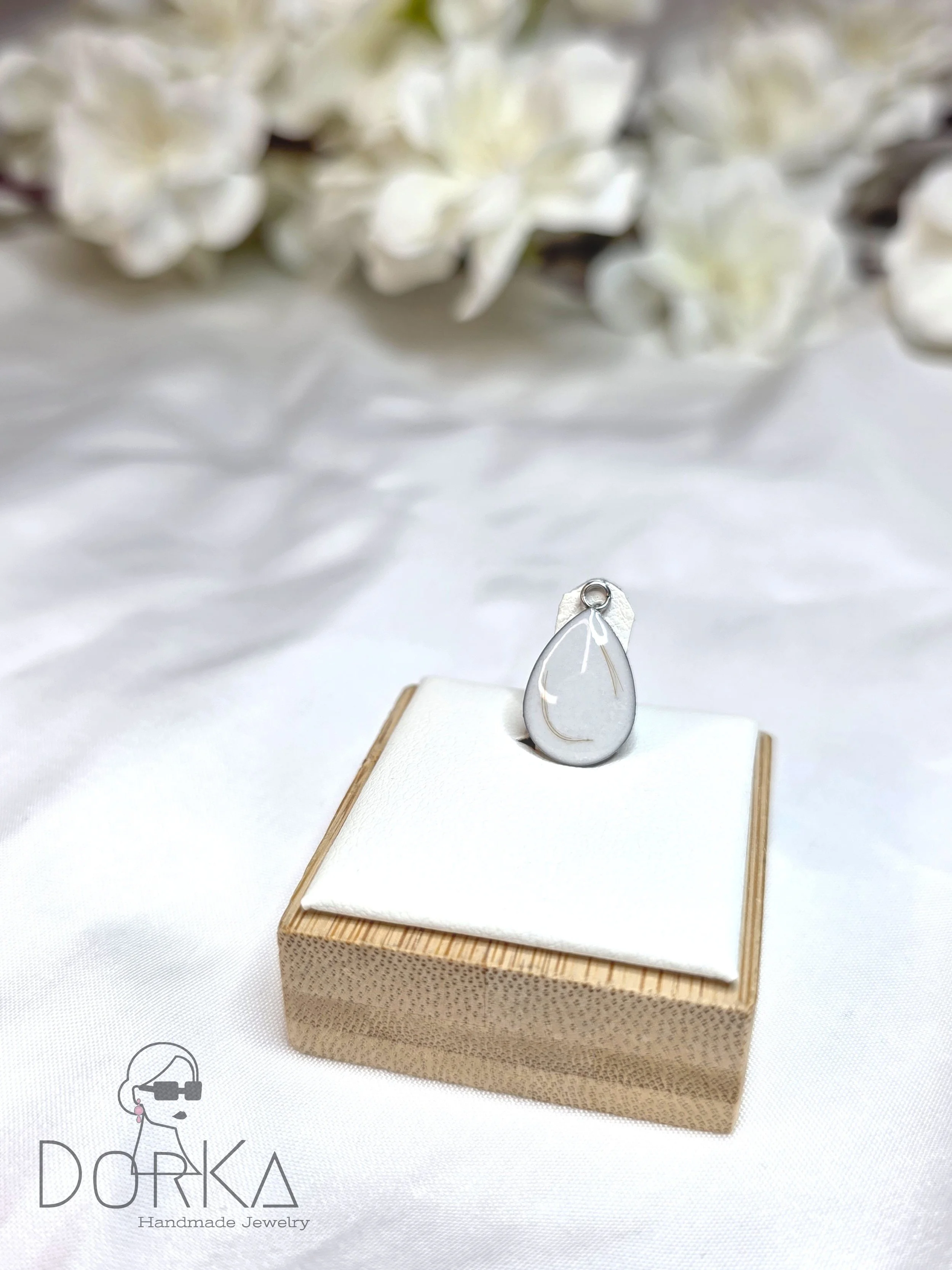 Silberanhänger in Tropfenform auf einer weißen Unterlage, auf einem Holzständer, mit weißen Blumen im Hintergrund.