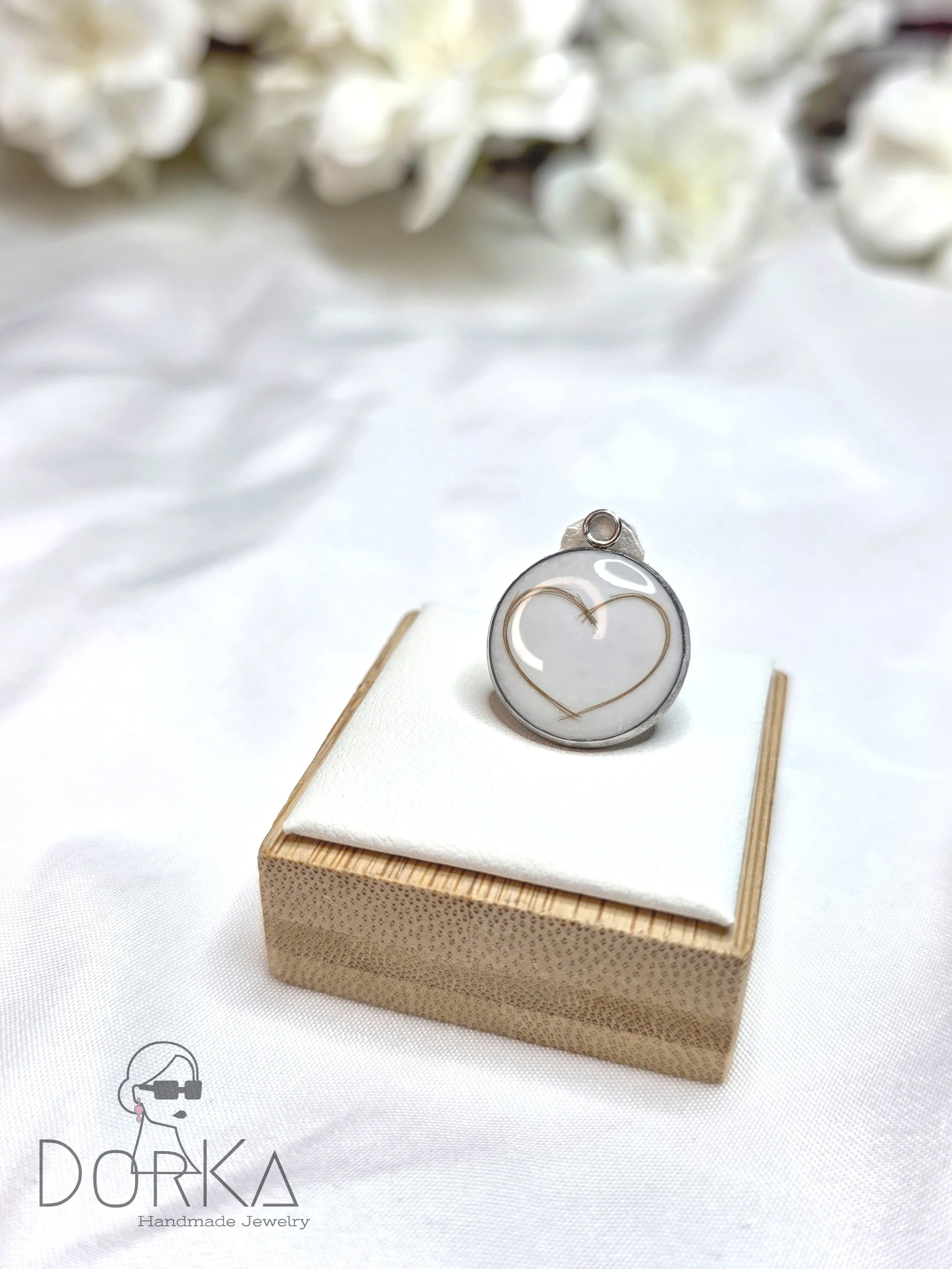 Kreisförmiger Anhänger mit einem goldenen Herz in der Mitte, präsentiert auf einer weißen Polsterung auf einem kleinen Holzkästchen, mit weißen Blumen im Hintergrund.