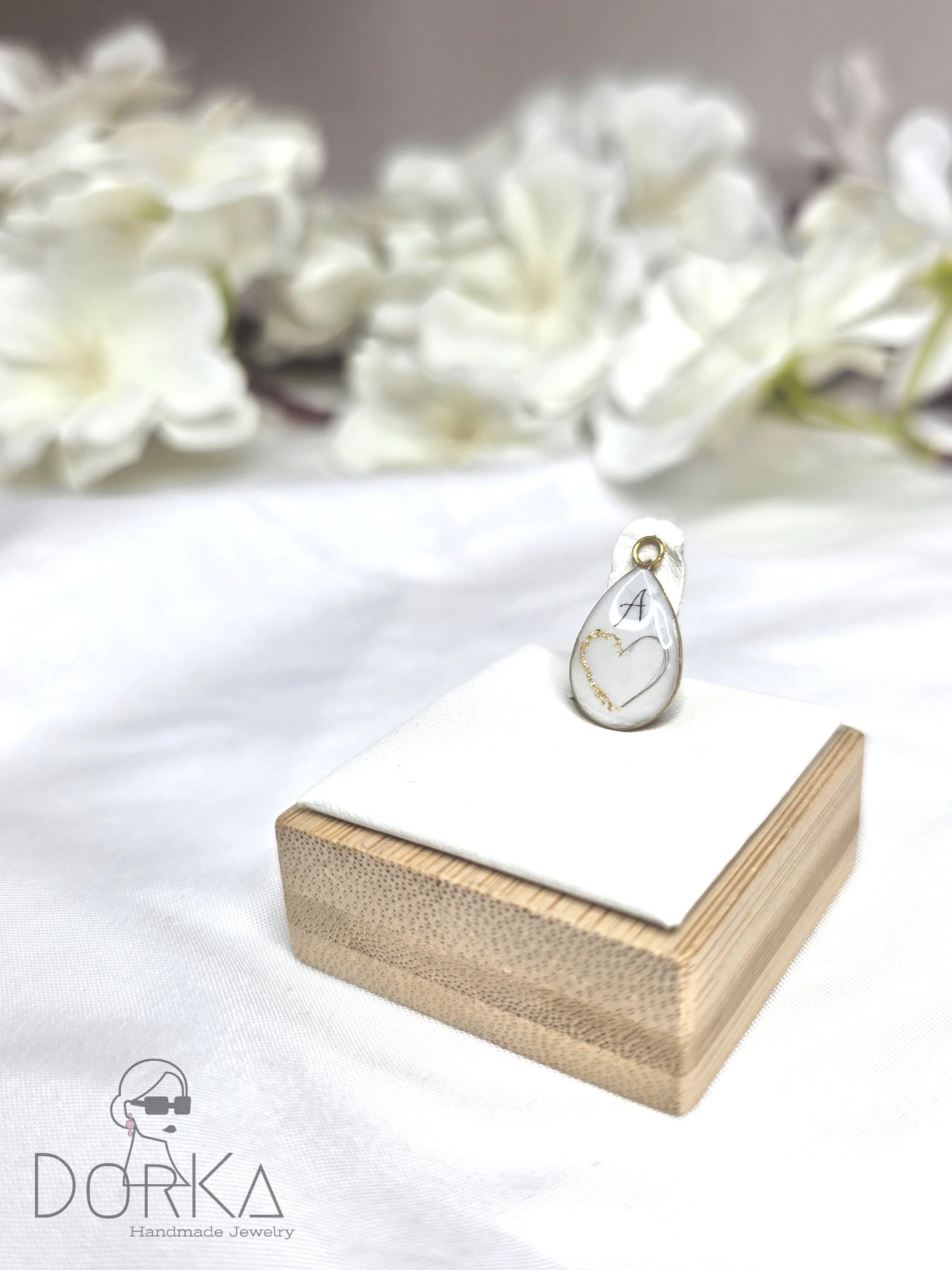 Ein handgefertigtes Schmuckstück in einer weißen Perle mit goldenen Details, auf einem weißen Kissen auf einem Holzboden, mit weißen Blumen im Hintergrund.