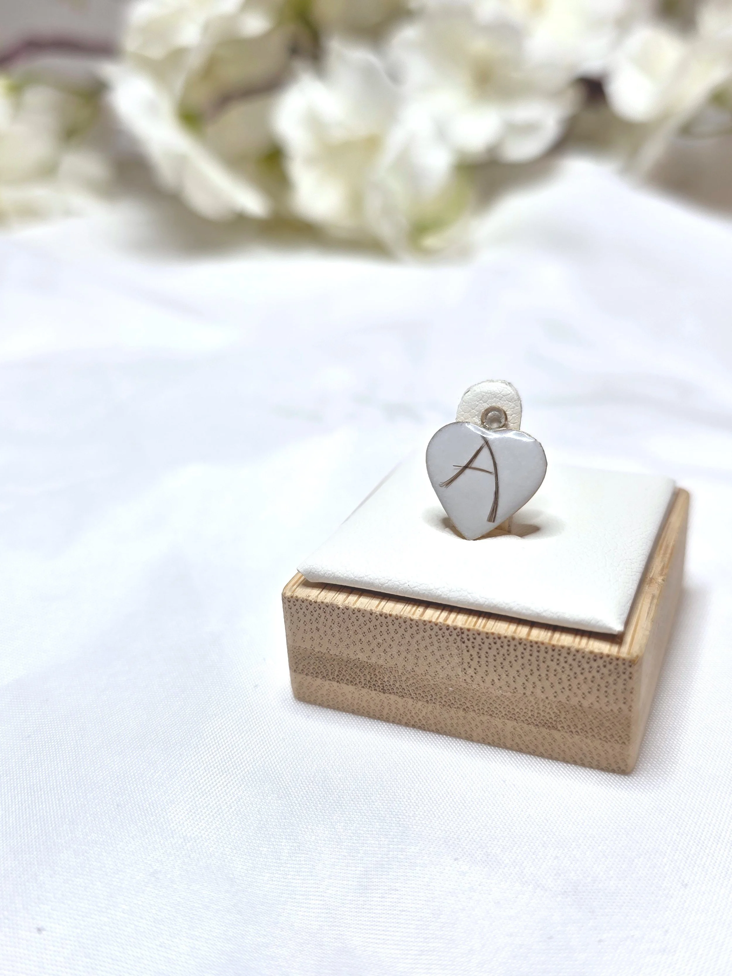 Ein kleiner Motivanhänger in Herzform mit einem Baum-Motiv, platziert auf einer kleinen Schmuckschatulle, vor einem weißen Hintergrund mit weißen Blumen im Hintergrund.