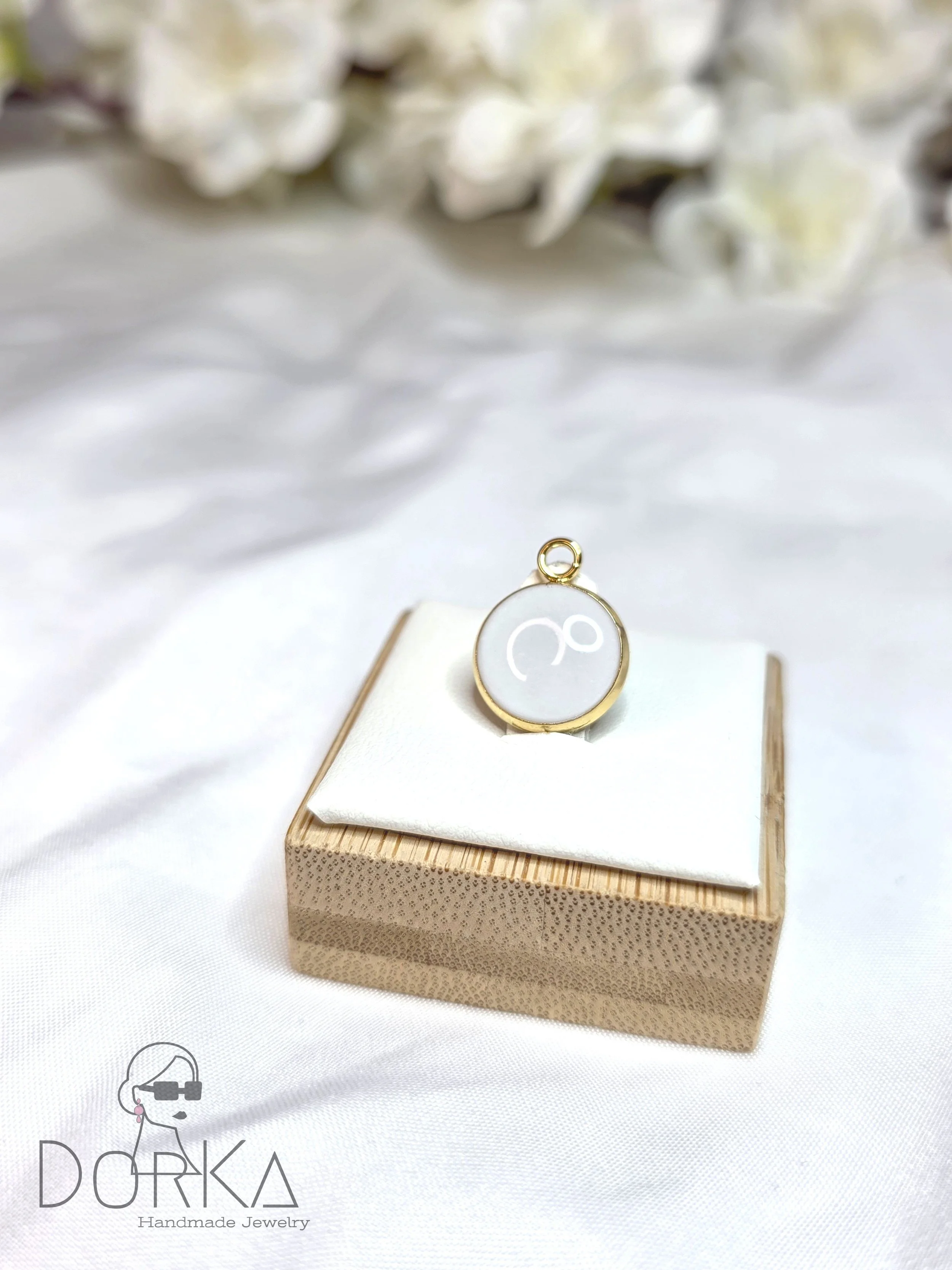 Eine runde handgemachte Halskette mit einem weißen Anhänger mit einem minimalistischen goldenen Knoten-Motiv, präsentiert auf einem weißen Kissen auf einem Holzkästchen, im Hintergrund weiße Blumen.