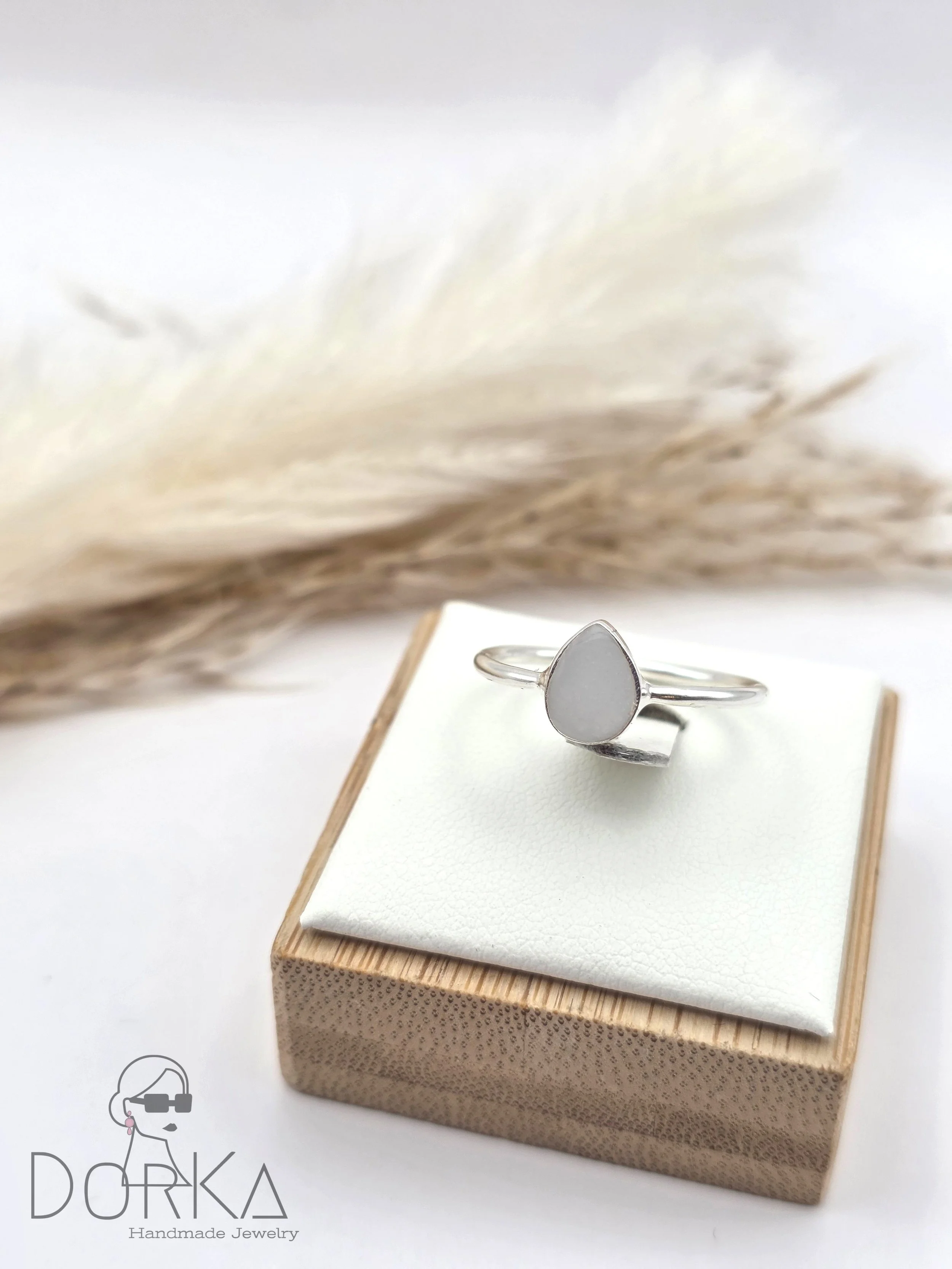 Silberarmreif mit weißem, tropfenförmigem Anhänger auf Schmuckschachtel, im Hintergrund getrocknete Weizenähren.