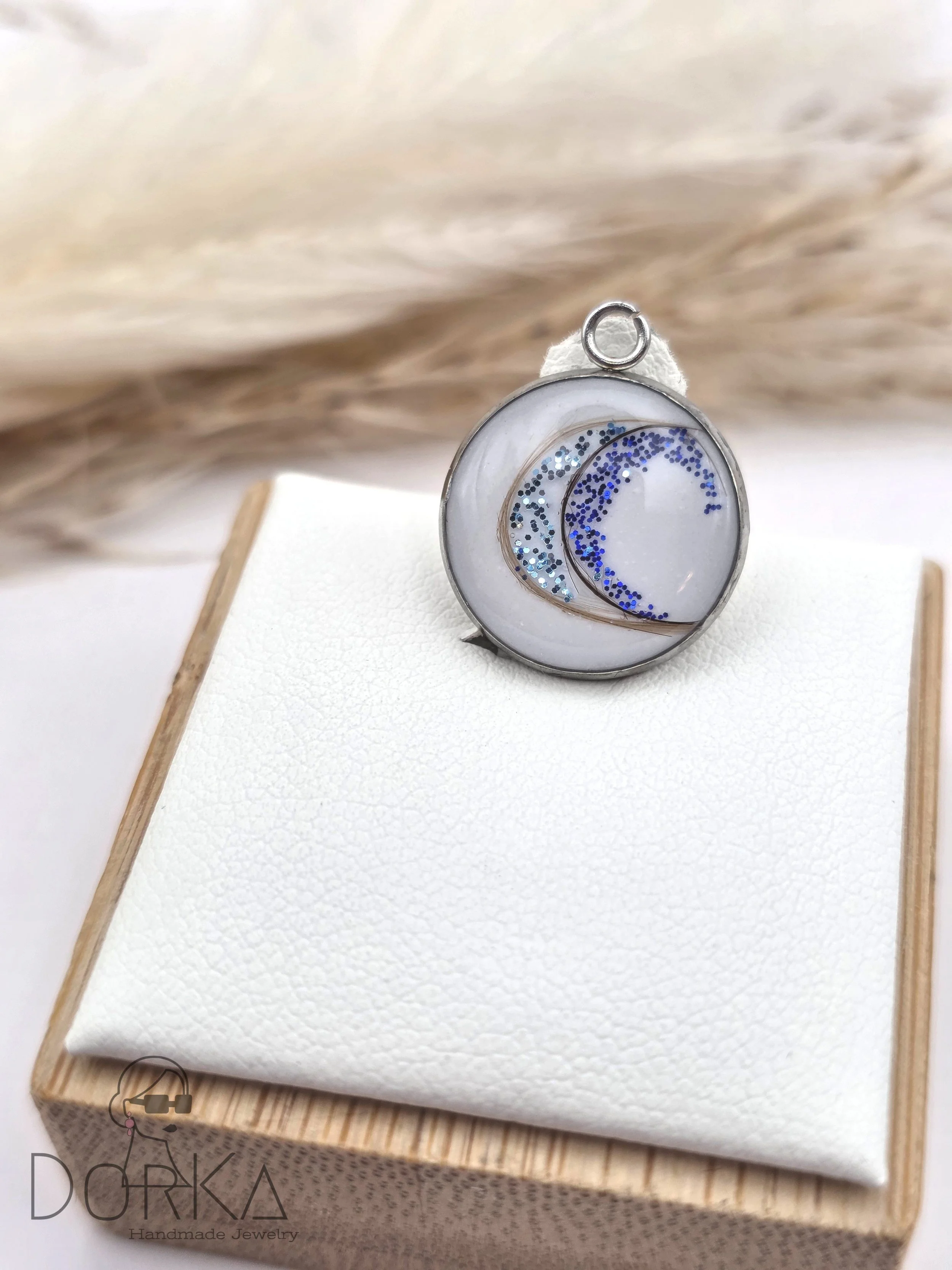 Ein runder Anhänger mit einem weißen Hintergrund und blauen Glitzerakzenten, präsentiert auf einer weißen Stoffunterlage auf einem Holzsockel.