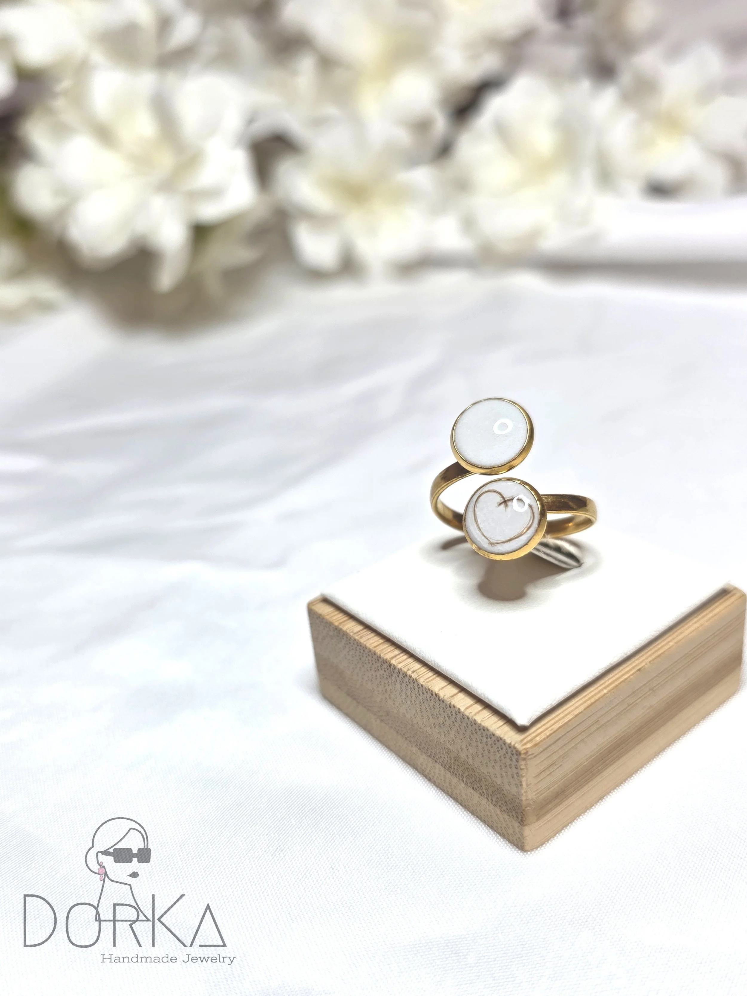 Goldener Ring mit zwei weißen runden Steinen auf einem weißen Podest, im Hintergrund weiße Blumen