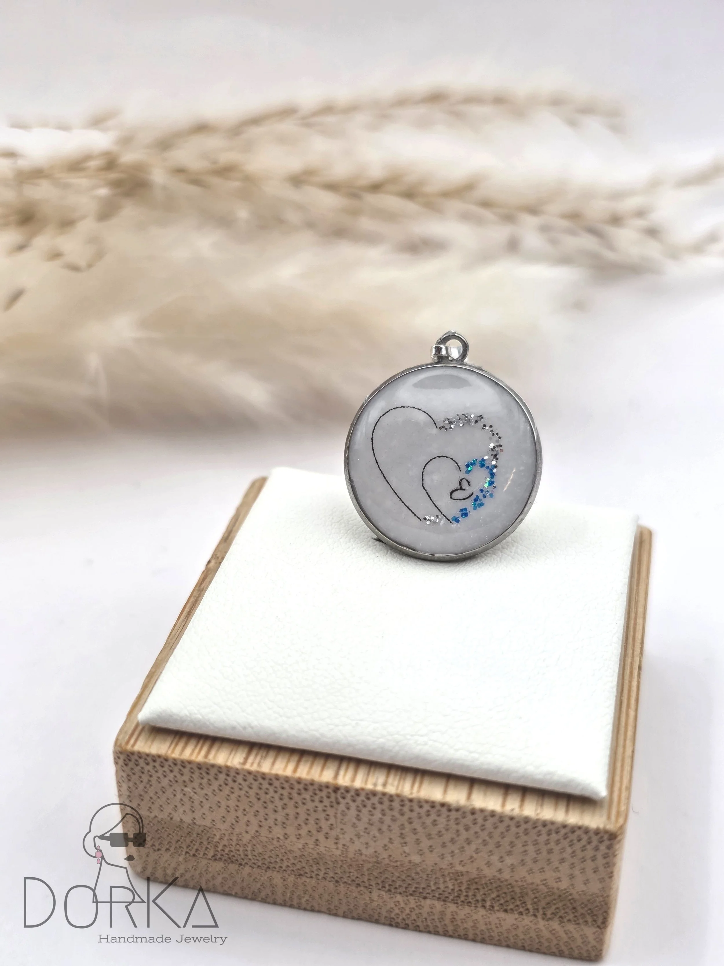 Rundes Schmuckstück mit zwei herzförmigen Linien und blauen und silberfarbenen Akzenten, auf einer weißen Stoffunterlage auf einem kleinen Holzkistchen, im Hintergrund getrocknetes Getreide.