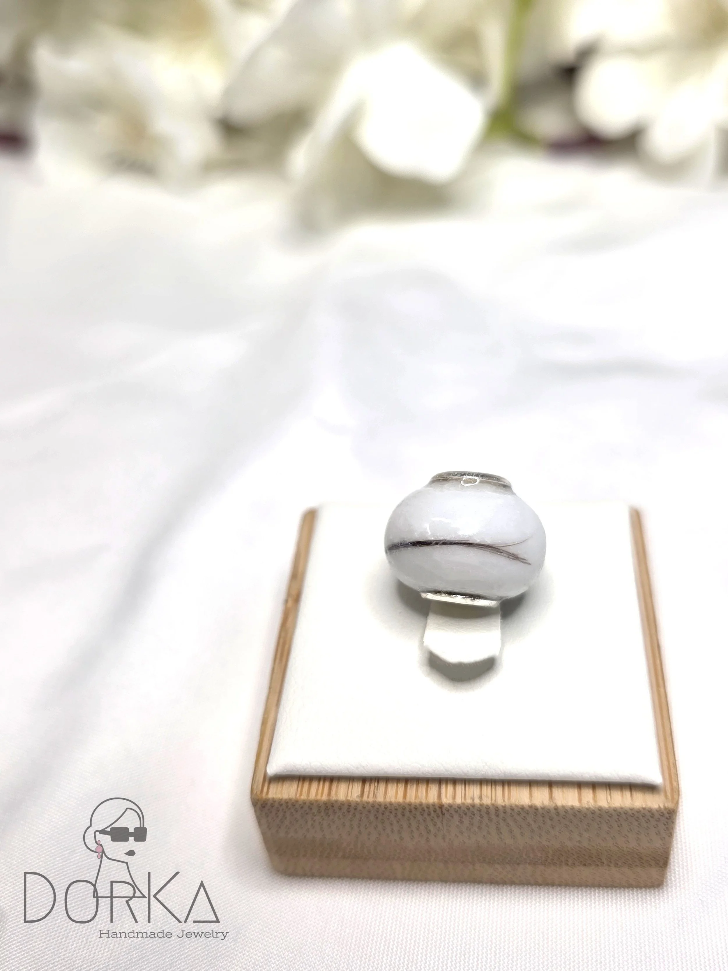 Ein kleines handgemachtes Schmuckstück auf einem weißen Kissen, präsentiert auf einem Holzsockel, mit dem Logo 'DORKA Handmade Jewelry' in der unteren linken Ecke.