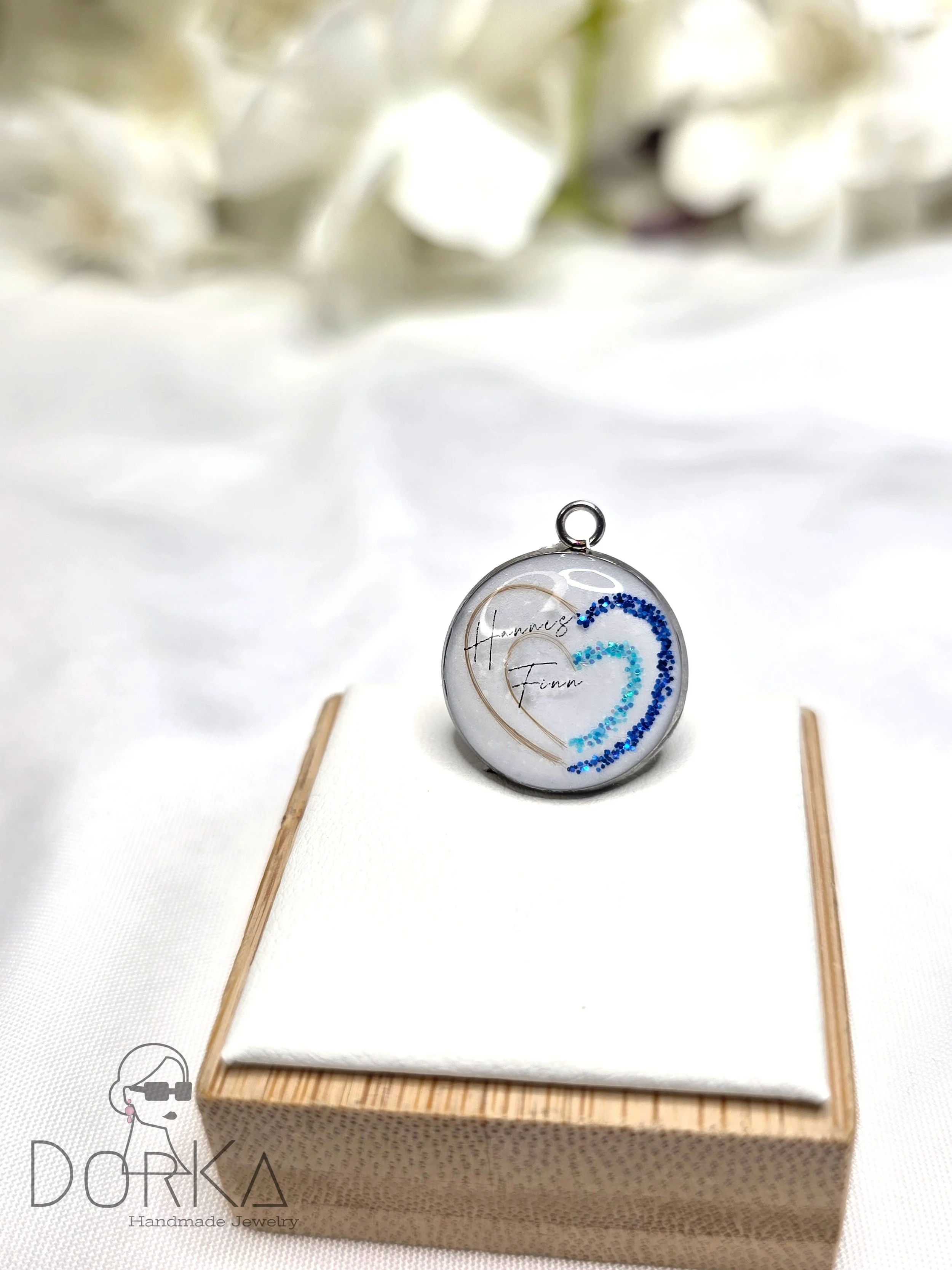 Ein runder Anhänger mit den Namen 'Hannes' und 'Finn' sowie blauen, geschwungenen Mustern, auf einem weißen Kissen in einer Holzschachtel, mit weißen Blumen im Hintergrund.