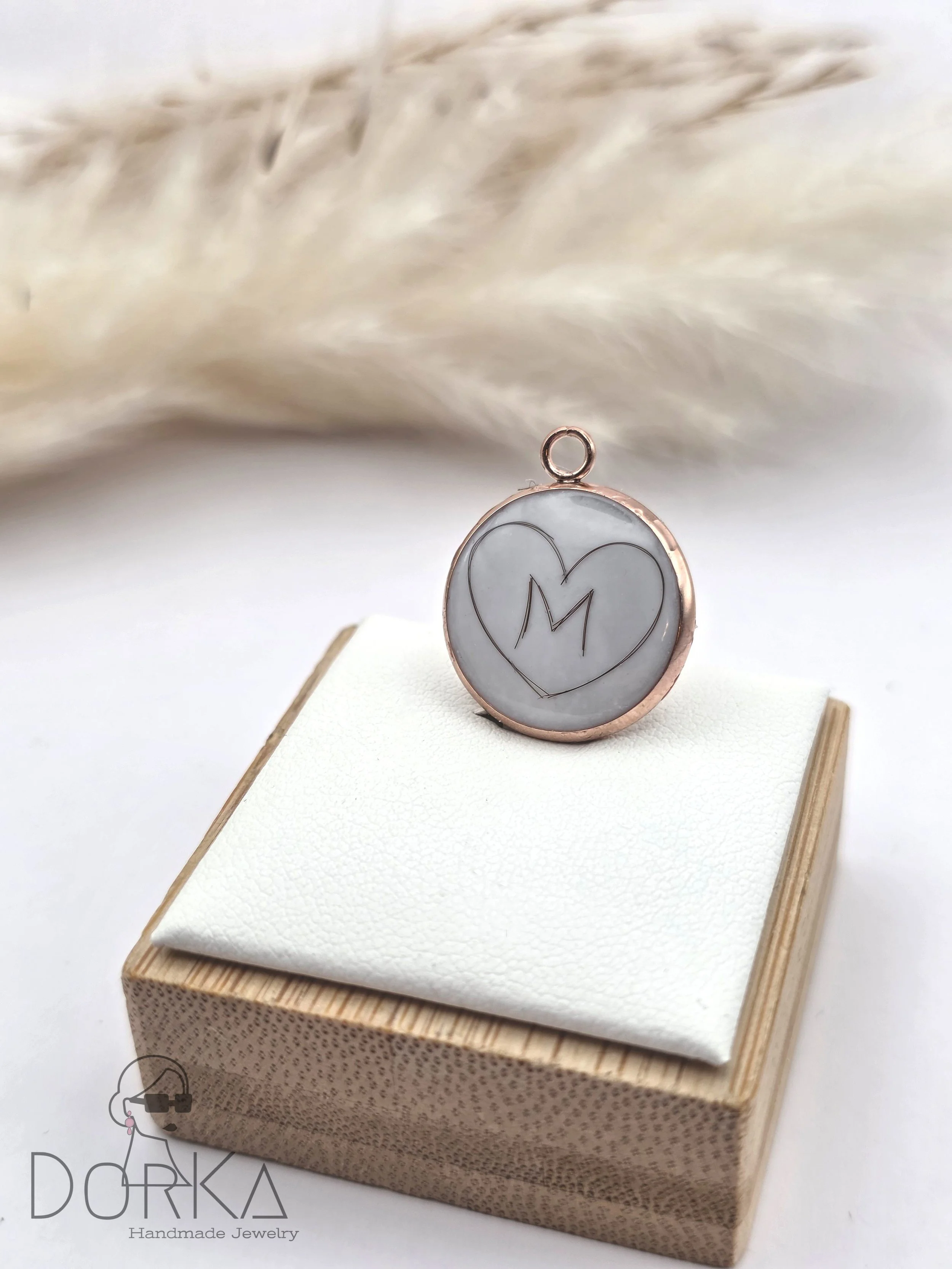 Runder Anhänger mit einer eingravierten Heart-Form und dem Buchstaben M, auf einem weißen Aufbewahrungsstück mit Holzkiste im Hintergrund.