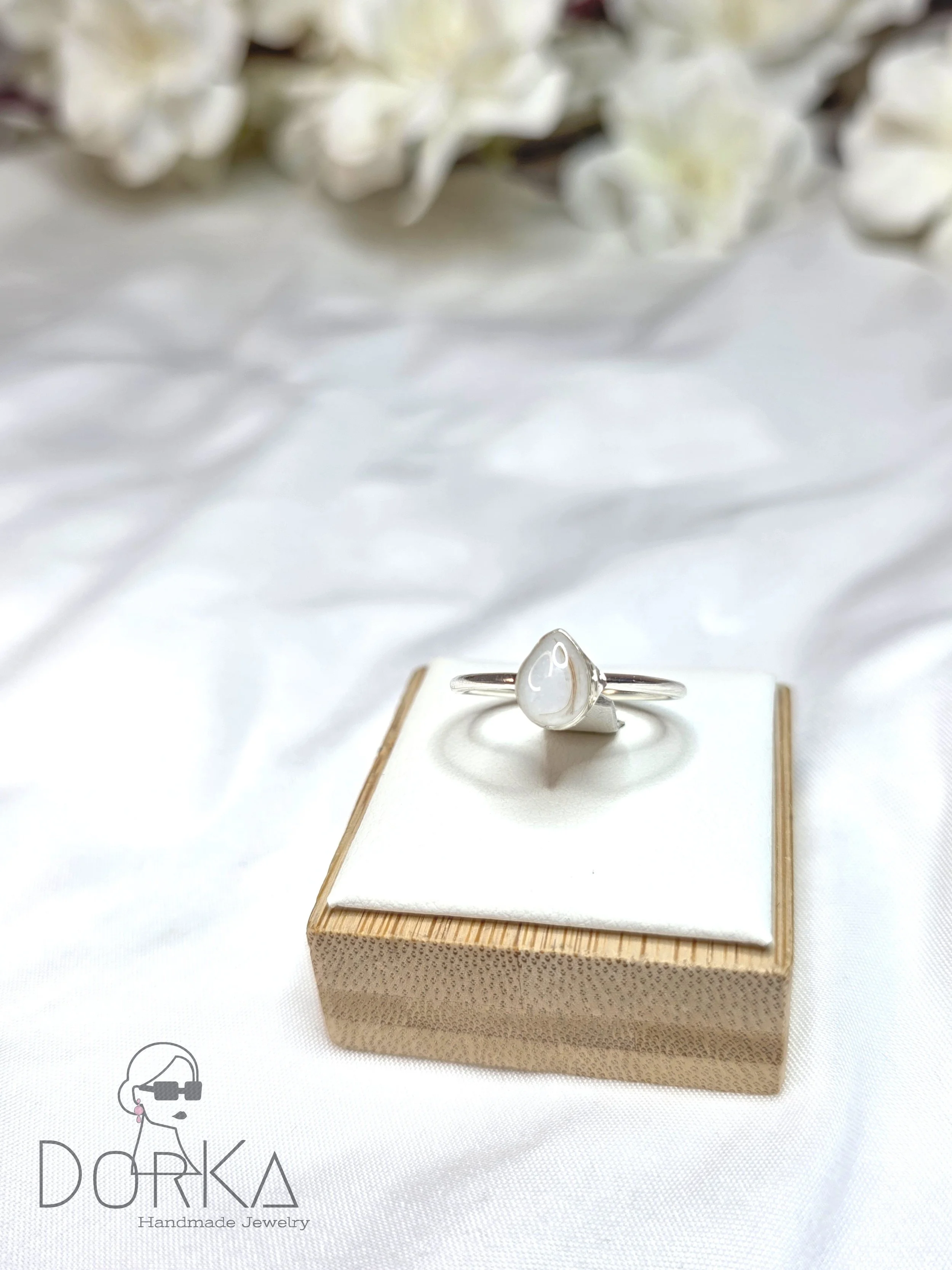 Silberring mit weißem, gestreiftem Stein auf einem kleinen weißen Kissen und Holzkästchen, im Hintergrund weiße Blumen.