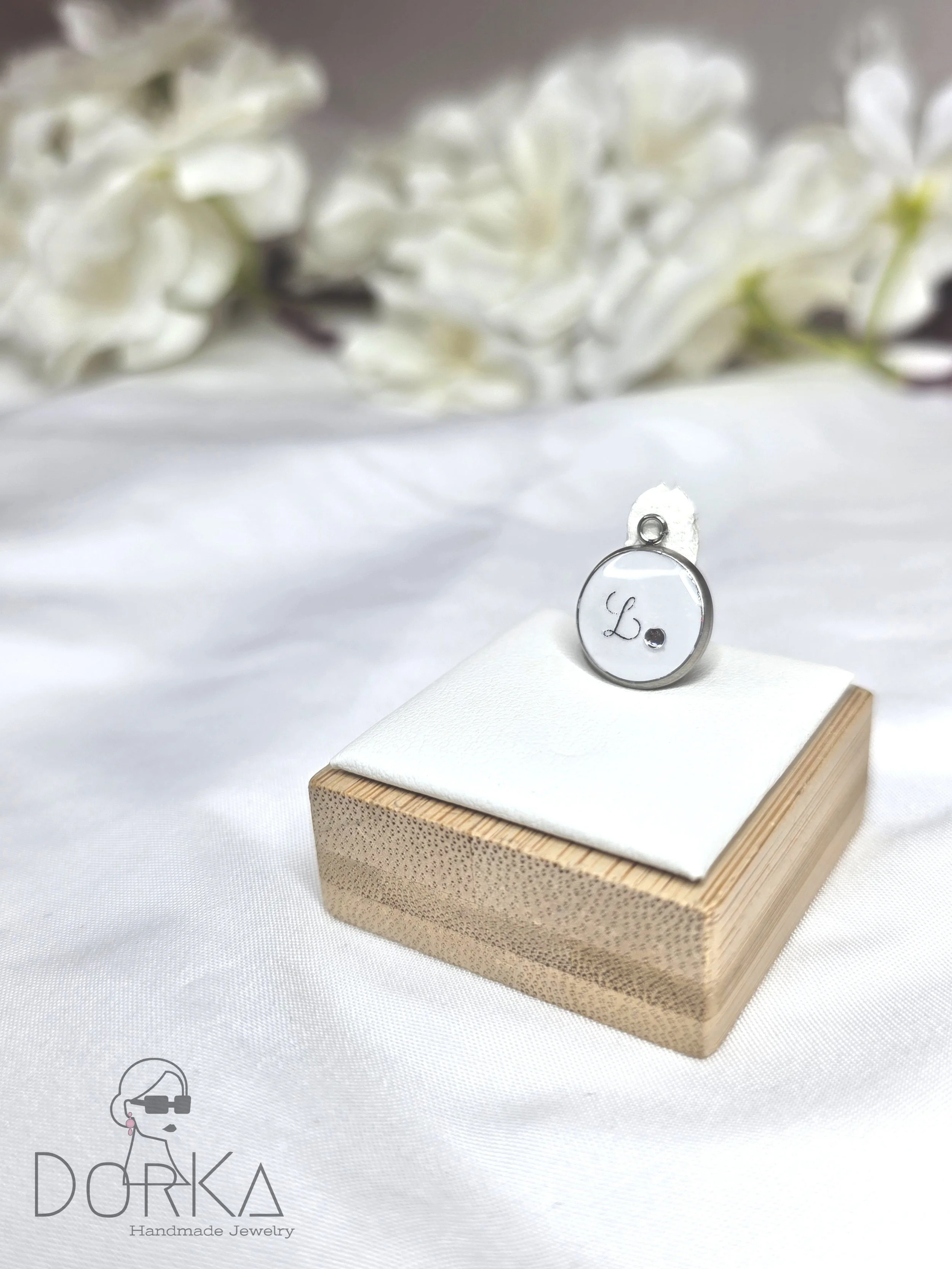 Eine kleine, runde, silberne Plakette mit dem Buchstaben ‚L‘ und einem kleinen Punkt, die auf einem weißen Stoffstück auf einem Holzblock liegt. Im Hintergrund sind weiße Blumen leicht unscharf. Am unteren linken Bildrand steht ein Logo mit der Aufsc