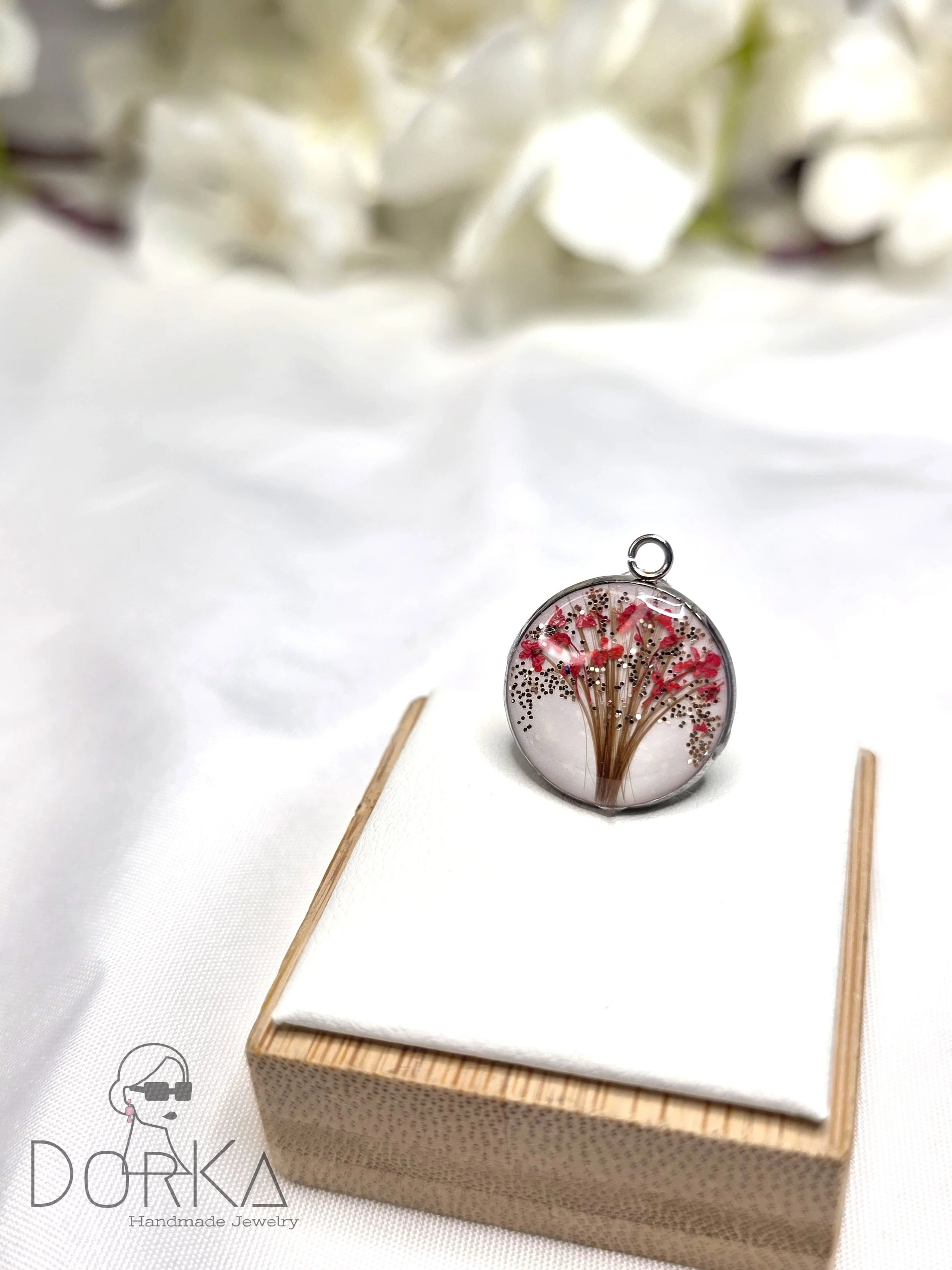 Runder Anhänger mit getrockneten Blumen in einer Silikonhülle, auf einem weißen Stoffrhombus auf einem kleinen Holzkästchen mit weißem Tuch, im Hintergrund weiße Blumen. Logo "DORKA Handmade Jewelry" im unteren linken Bereich.