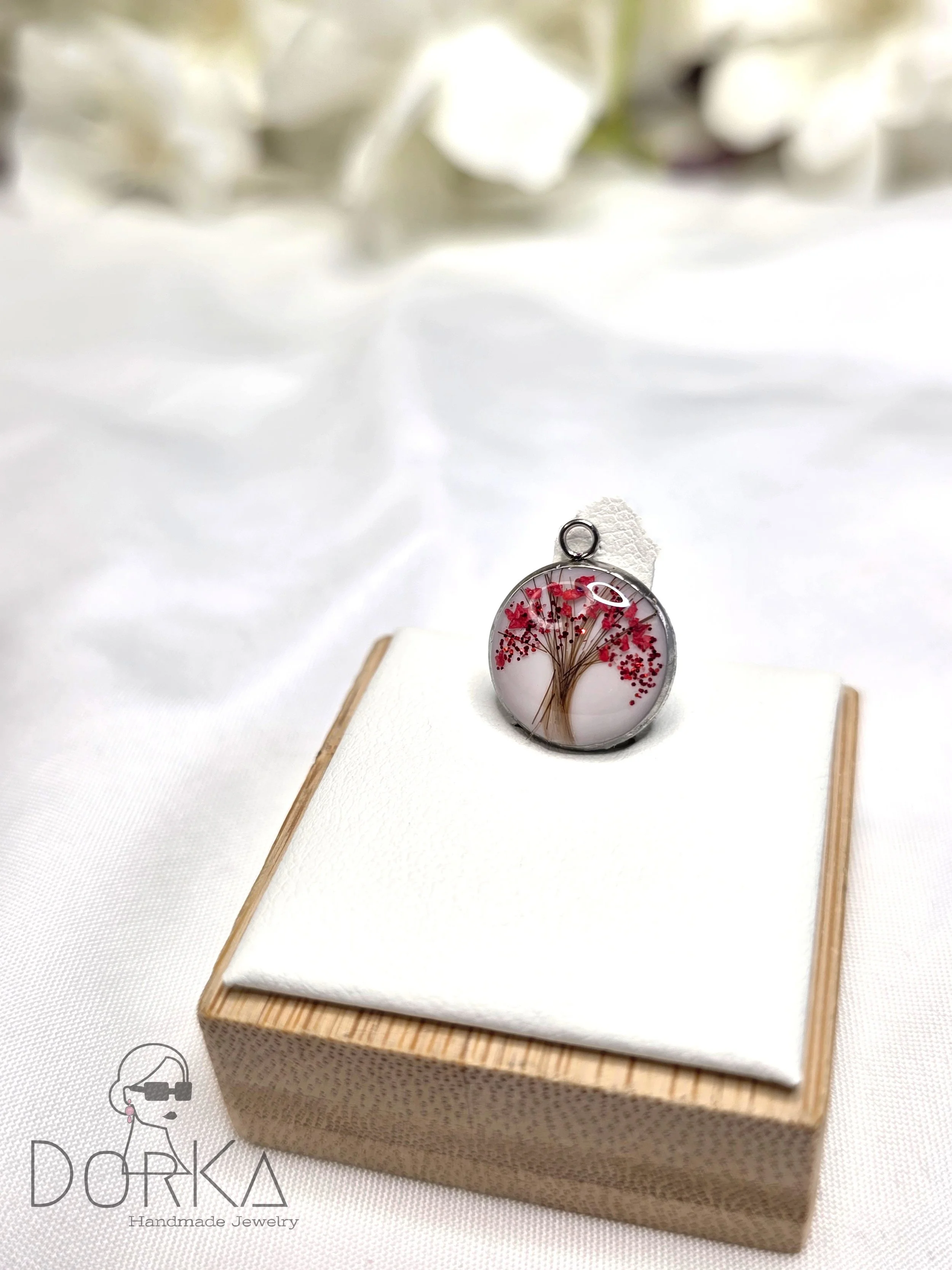 Ein runder Anhänger mit einem Baum-Design, das rote Blätter und braune Zweige zeigt, auf einem weißen Kissen auf einem Holzsockel. Im Hintergrund weiße Blumen.