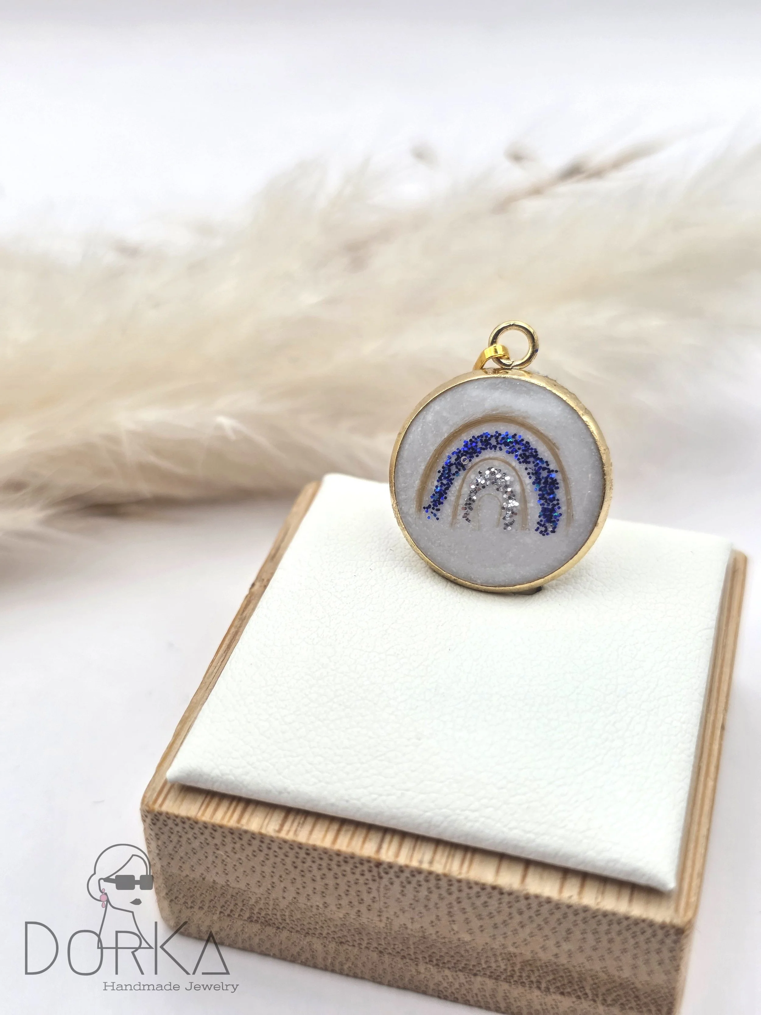 Ring mit Regenbogen-Design aus Glitzer auf weißem Hintergrund, auf einem weißen Stoffstück auf einem Holzsockel, mit pampasartigen Pflanzen im Hintergrund.