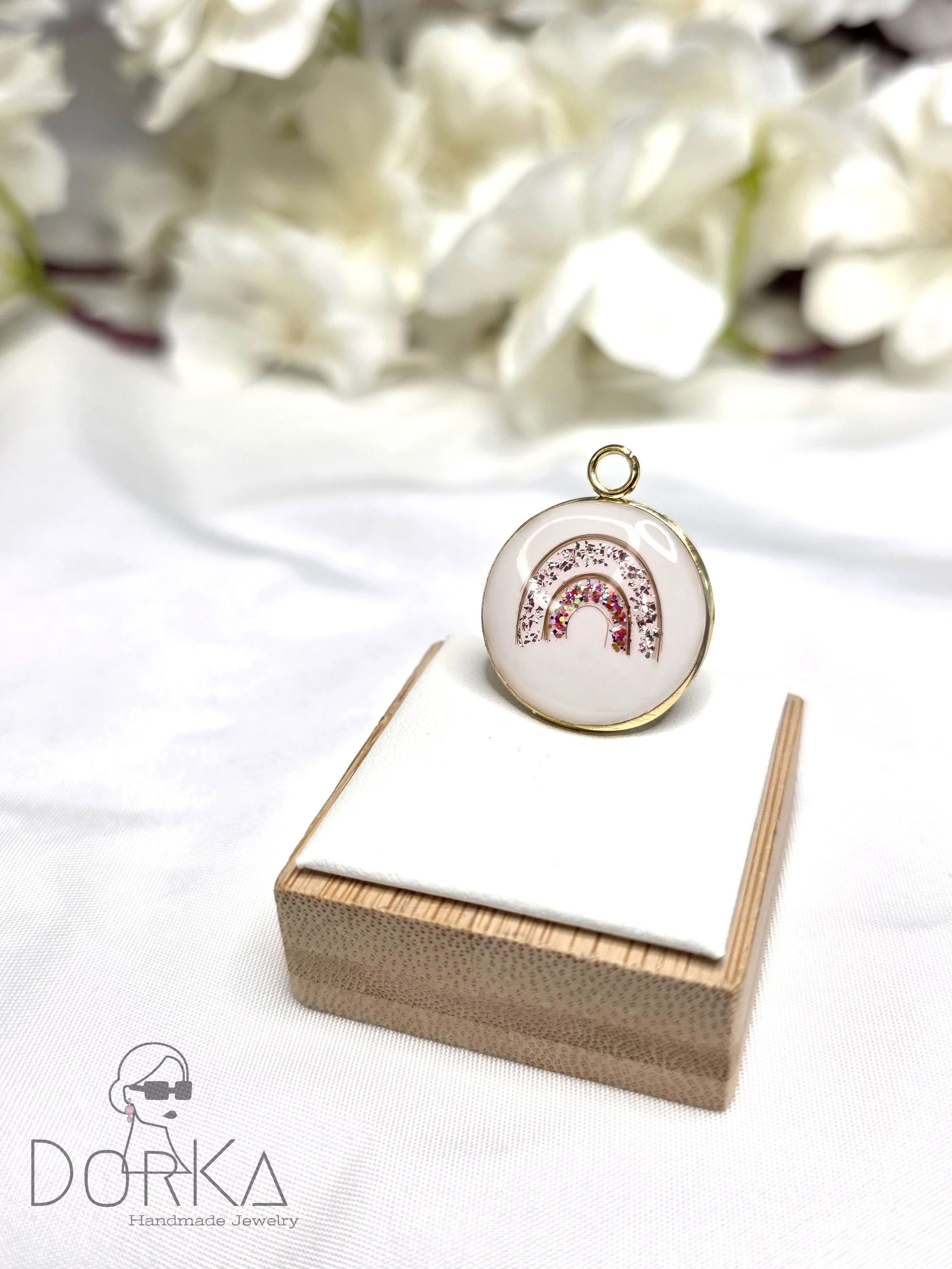 Schöner runder Anhänger mit Regenbogen-Design, dekoriert mit pinkfarbenen, silbernen und kristallfarbenen Steinen, auf einem weißen Stoffkissen und einem kleinen Holzkästchen, im Hintergrund weiße Blumen.