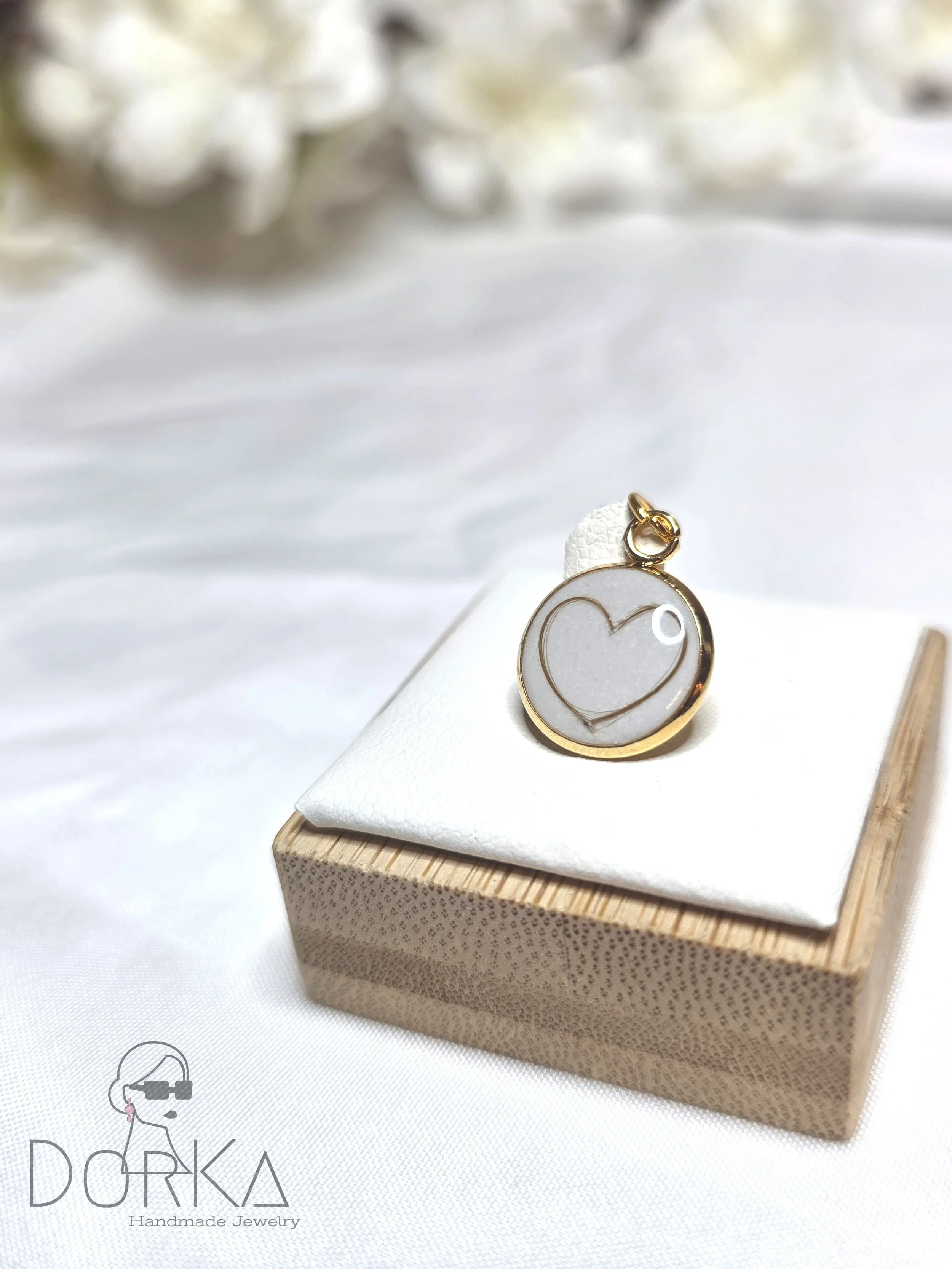 Rundes Schmuckstück mit Herz-Design, in Goldfassung, auf weißer Unterlage auf einem kleinen Holzkistchen. Hintergrund unscharf mit weißen Blumen.