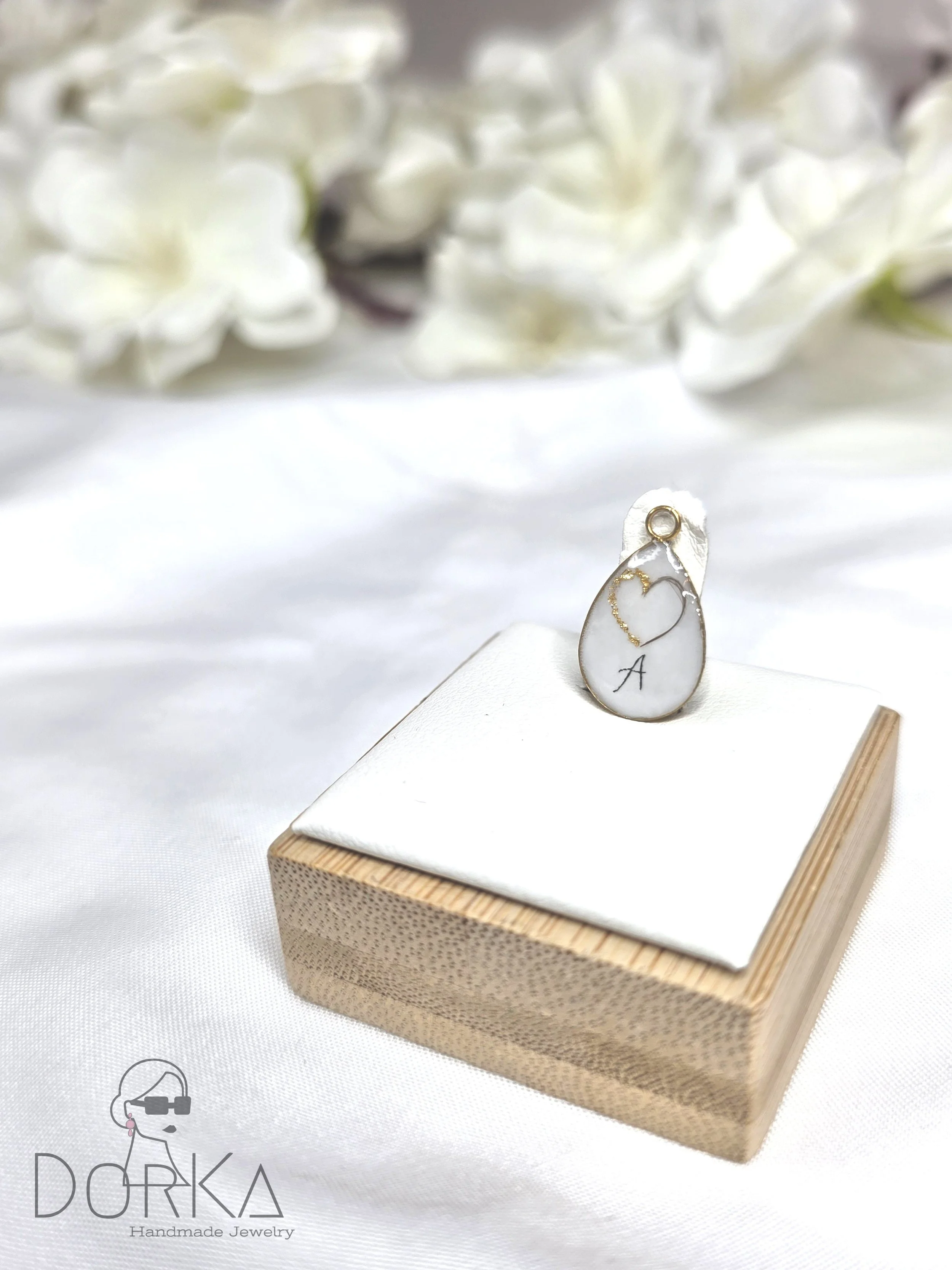 Handgefertigtes Schmuckstück auf einem weißen Stoffkissen, präsentiert auf einem Holzsockel, mit weißen Blumen im Hintergrund.