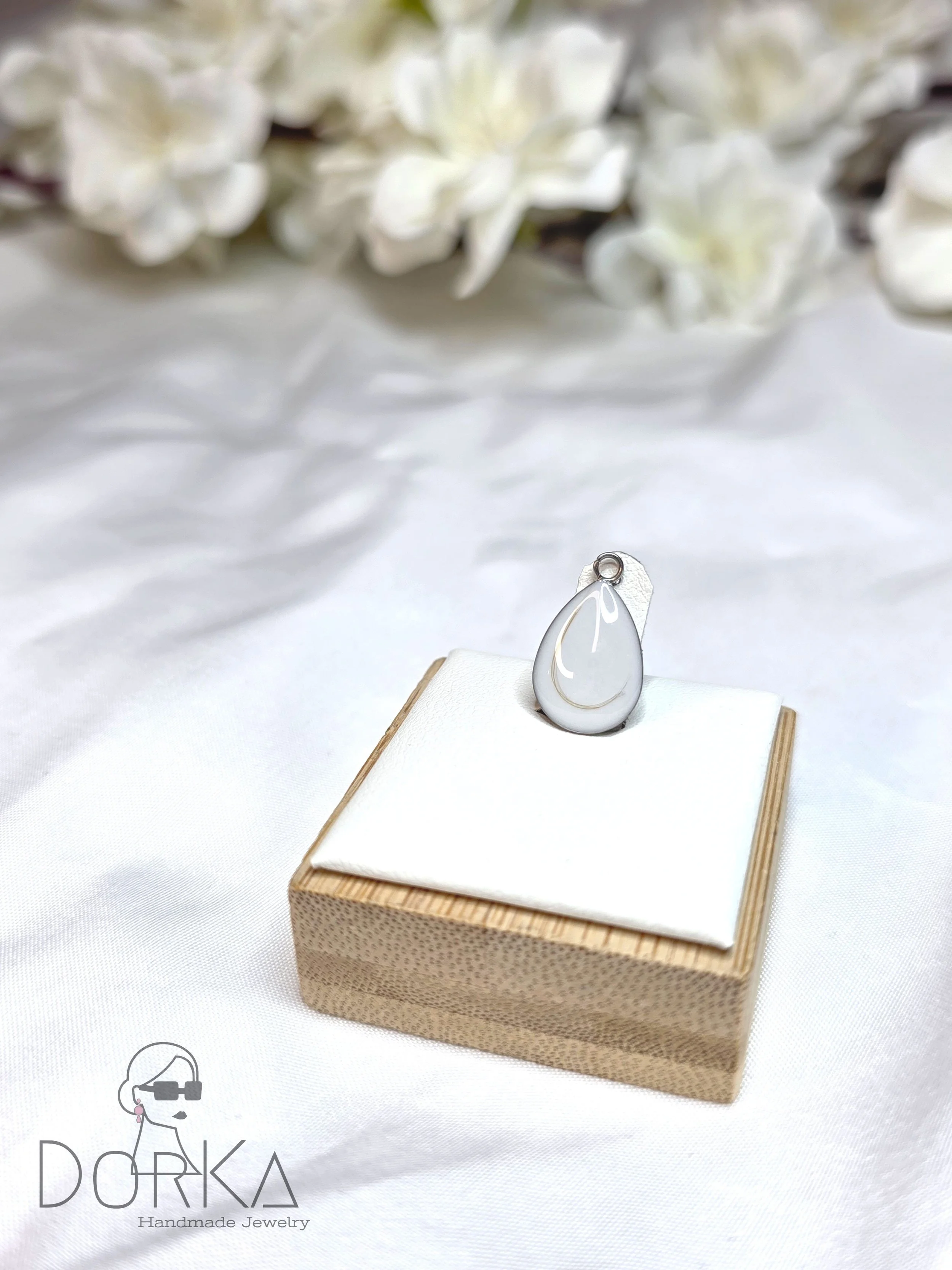 Ein silberner Anhänger in Form eines Tropfens auf einer weißen Stoffunterlage, auf einem kleinen Holzkästchen, mit weißen Blumen im Hintergrund.